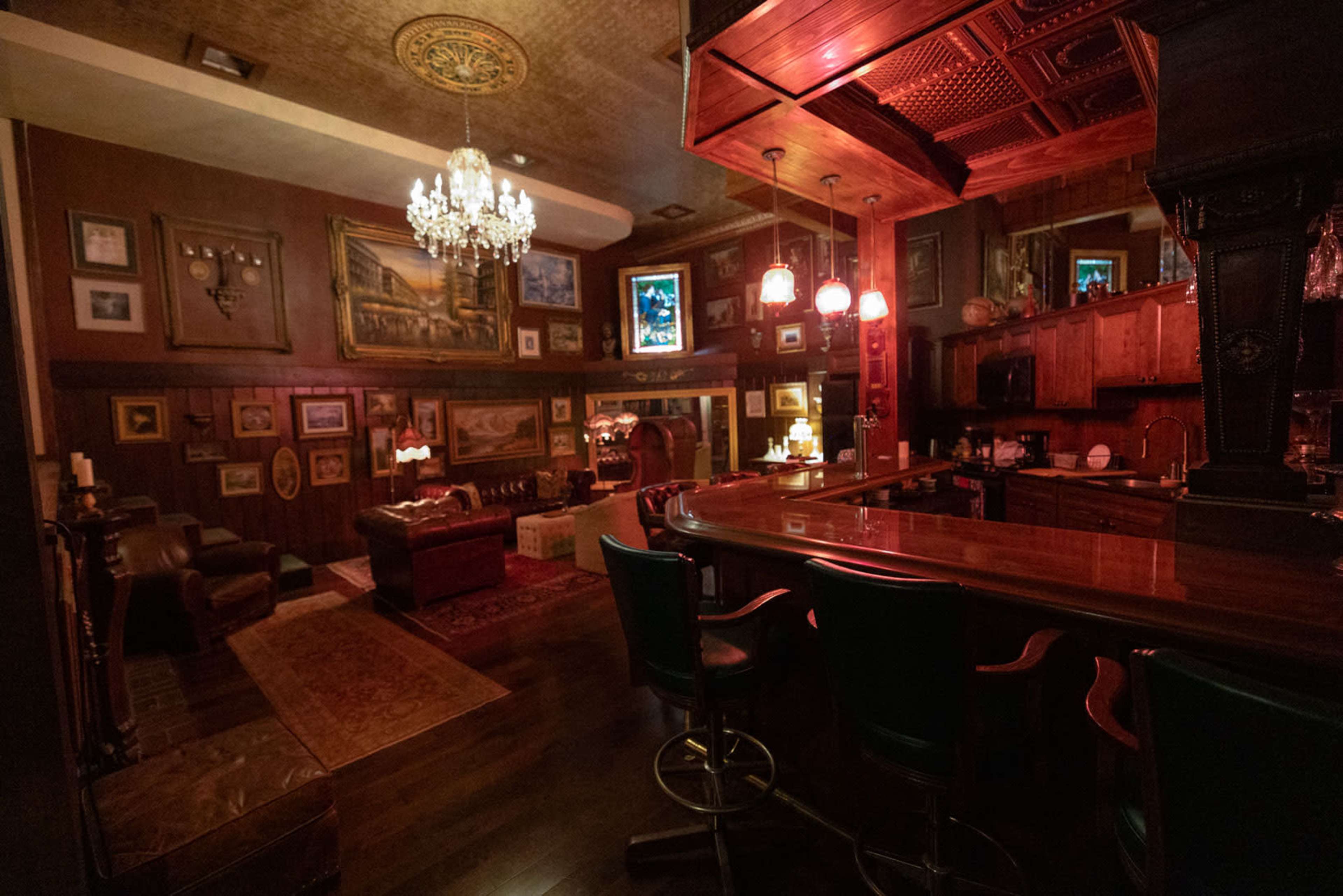 The image depicts a dimly lit, elegantly decorated bar area with wooden furnishings, a chandelier, and numerous framed artworks on the walls.