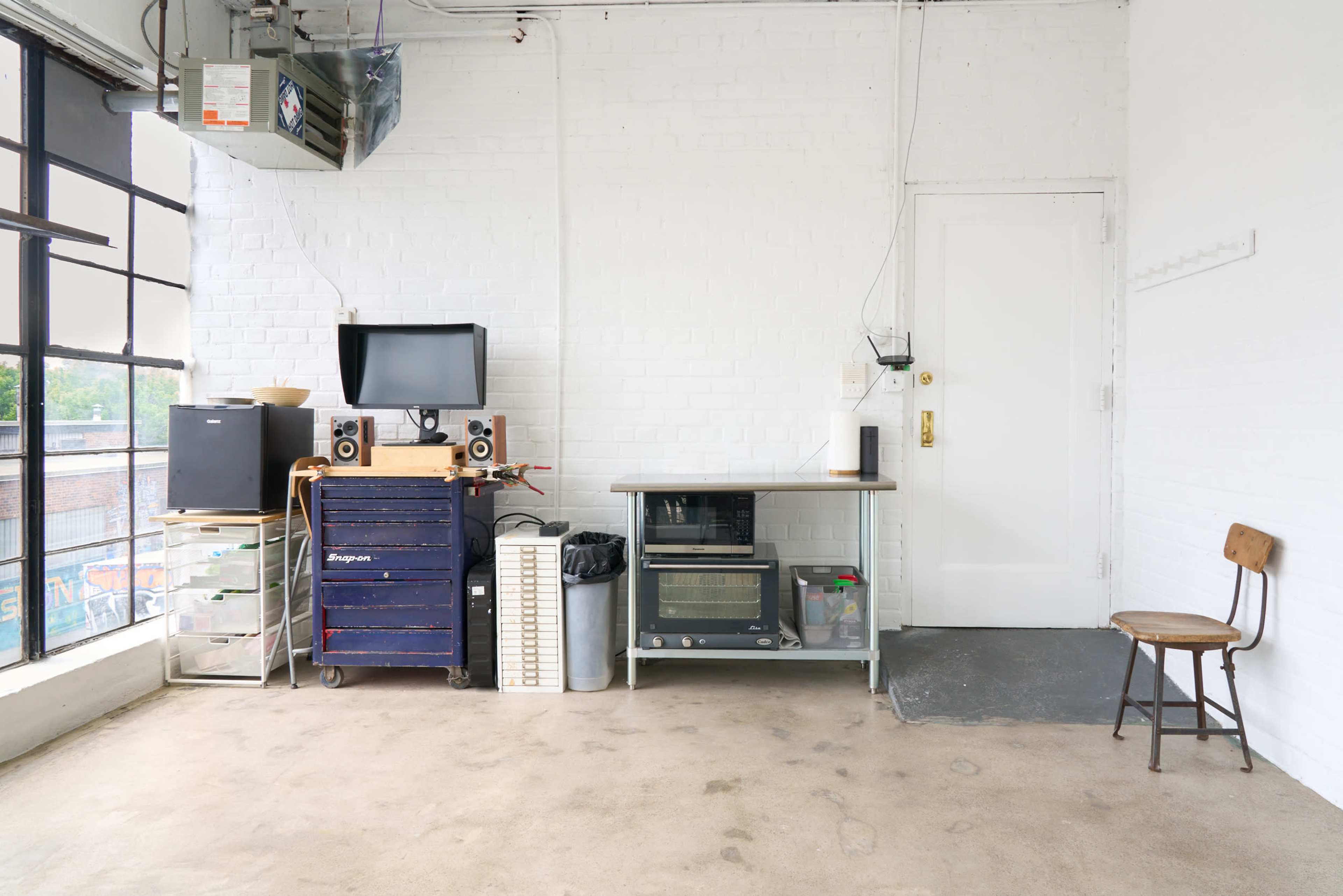 The image shows a sparse, brightly lit room with a white brick wall, featuring a small desk with an old television, speakers, and a chair against one side.