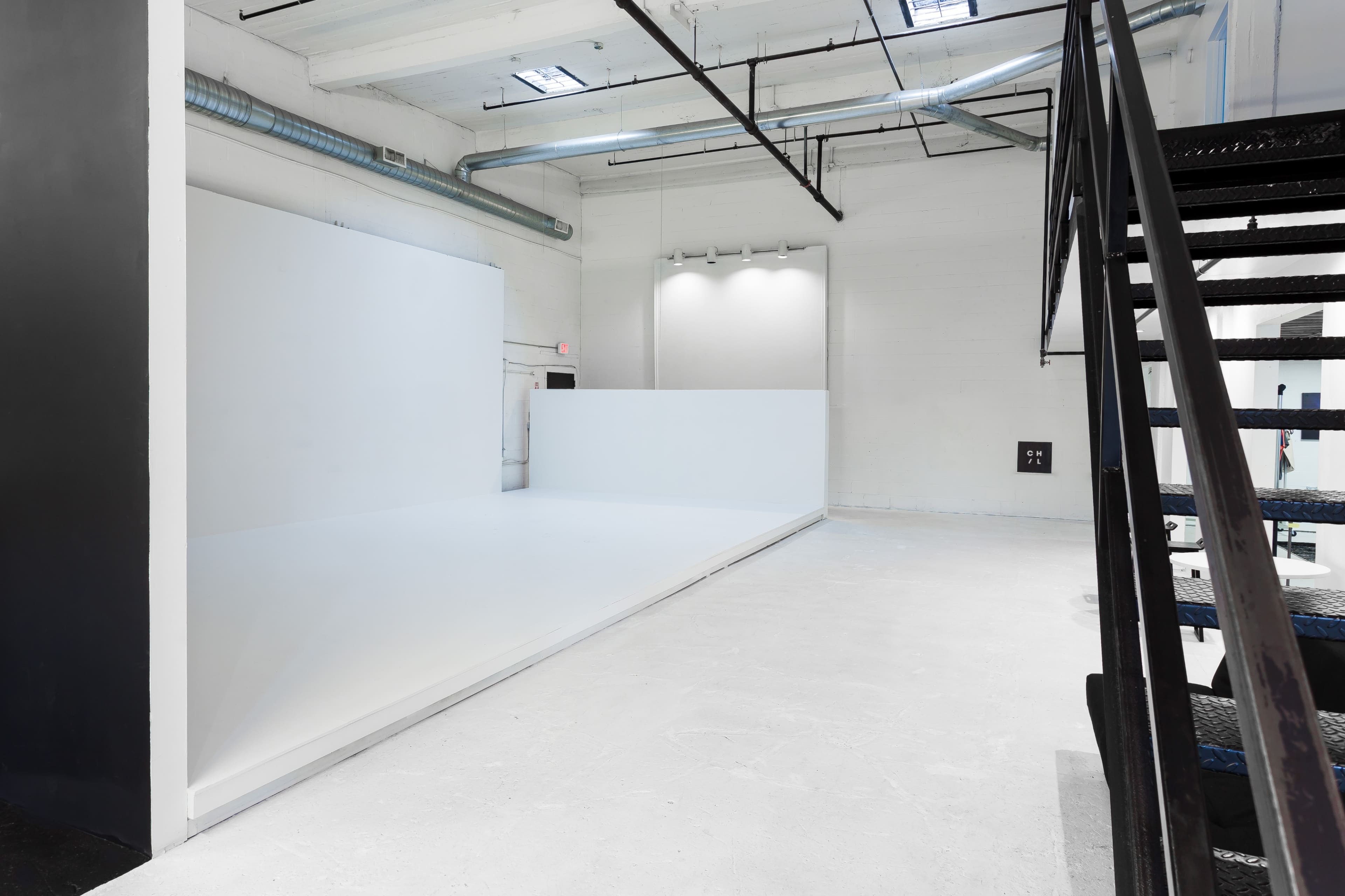 The image shows a minimalistic white studio space with a raised platform and a staircase in the foreground.