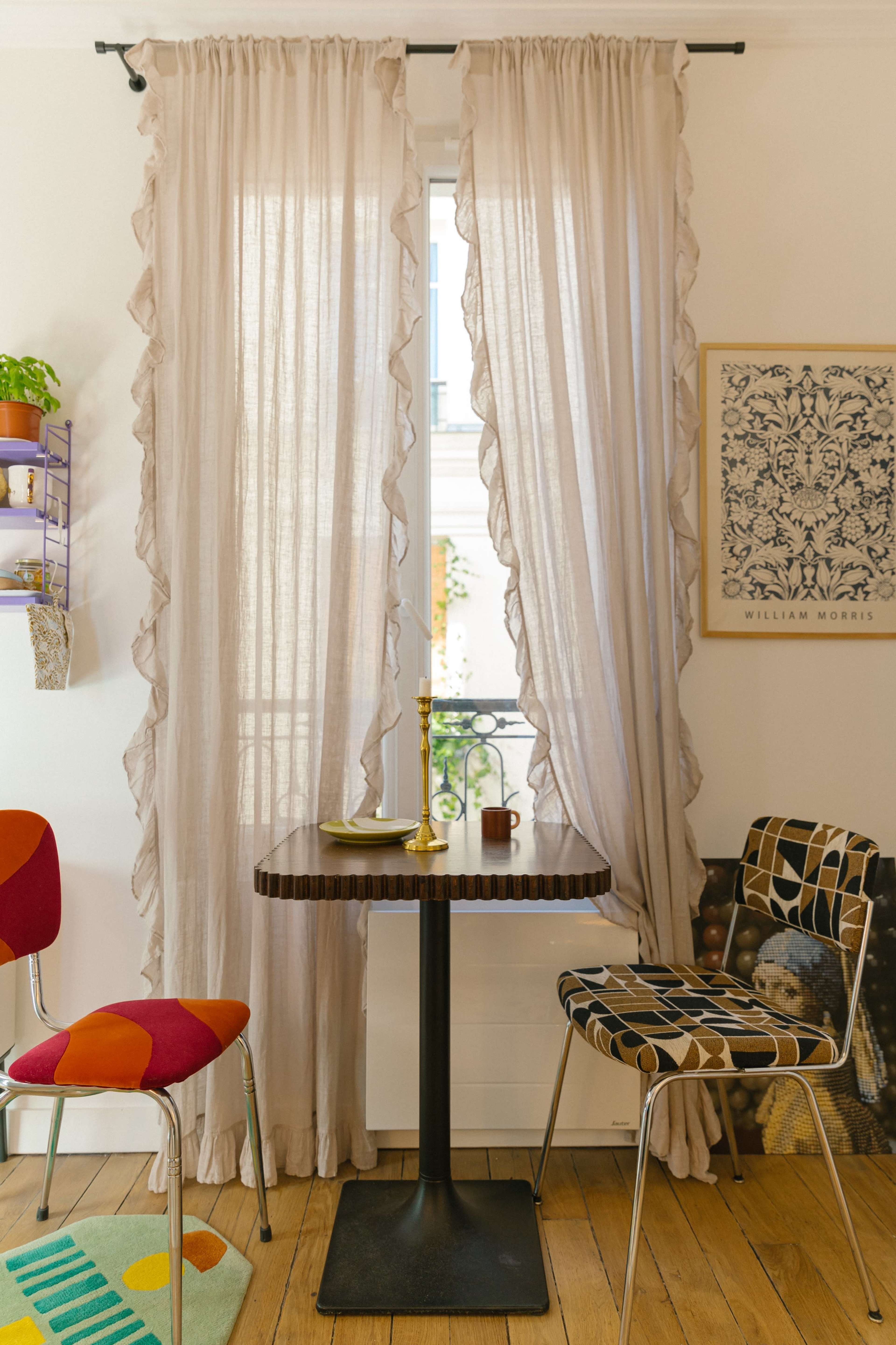 A small dining table with a candle stands between two chairs in a well-lit room, framed by sheer curtains and decorated with art on the wall.