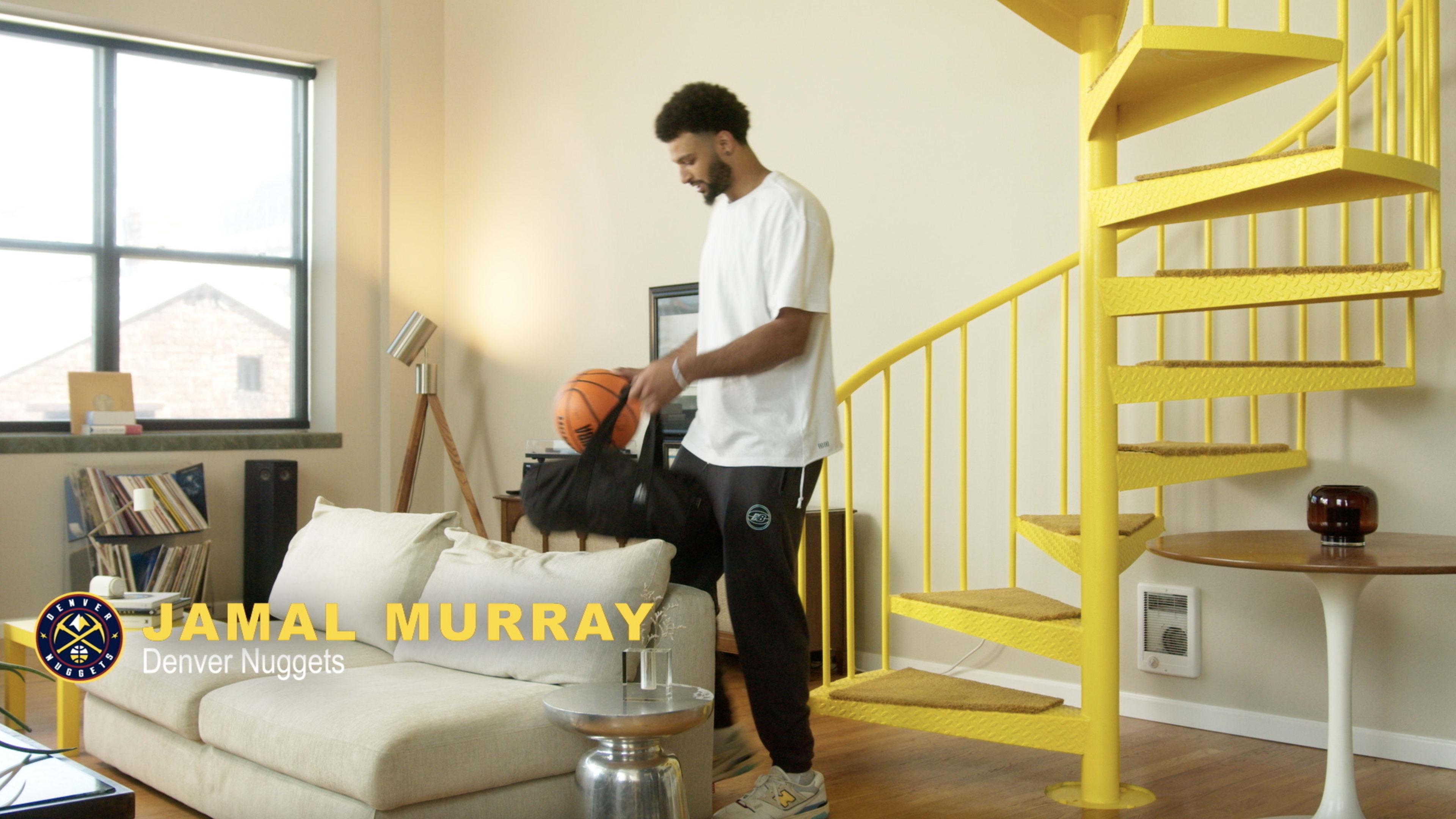 A man in a white t-shirt and sweatpants holds a basketball while standing next to a sofa in a brightly lit room with a spiral staircase.