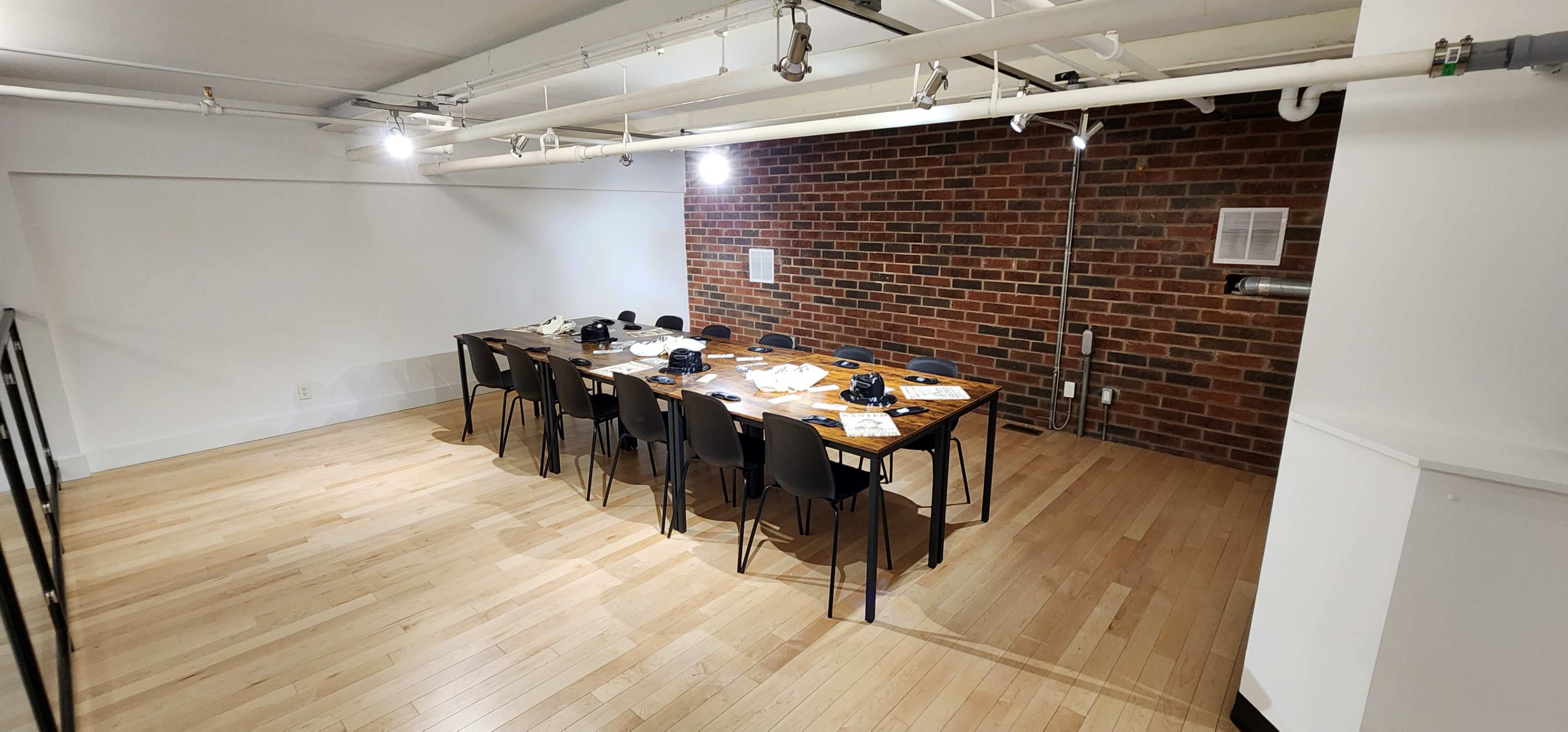 A long wooden table with black chairs is set up in a room featuring a brick wall and wooden flooring.