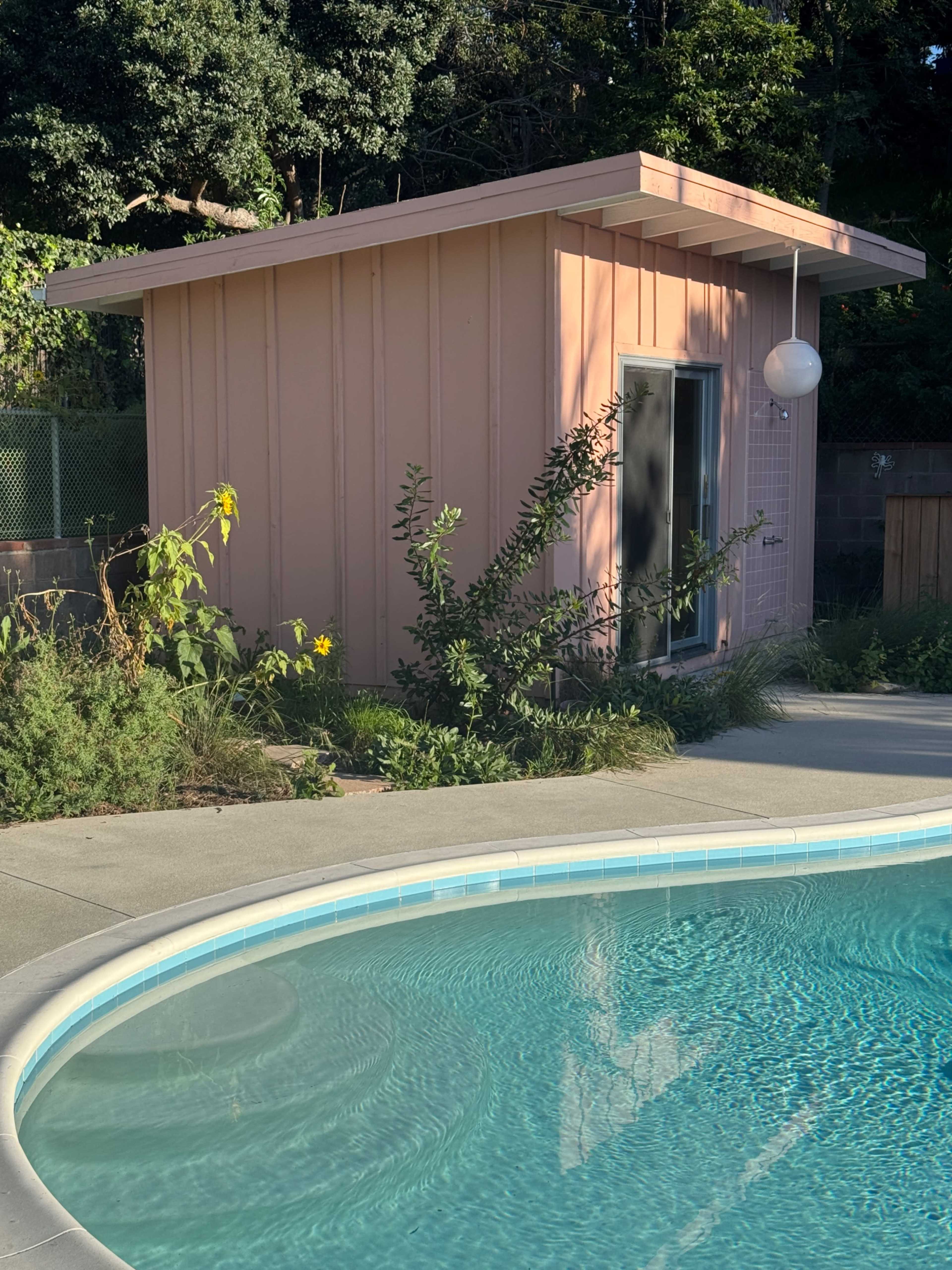 A pink shed is situated near a circular swimming pool surrounded by greenery and flowers.