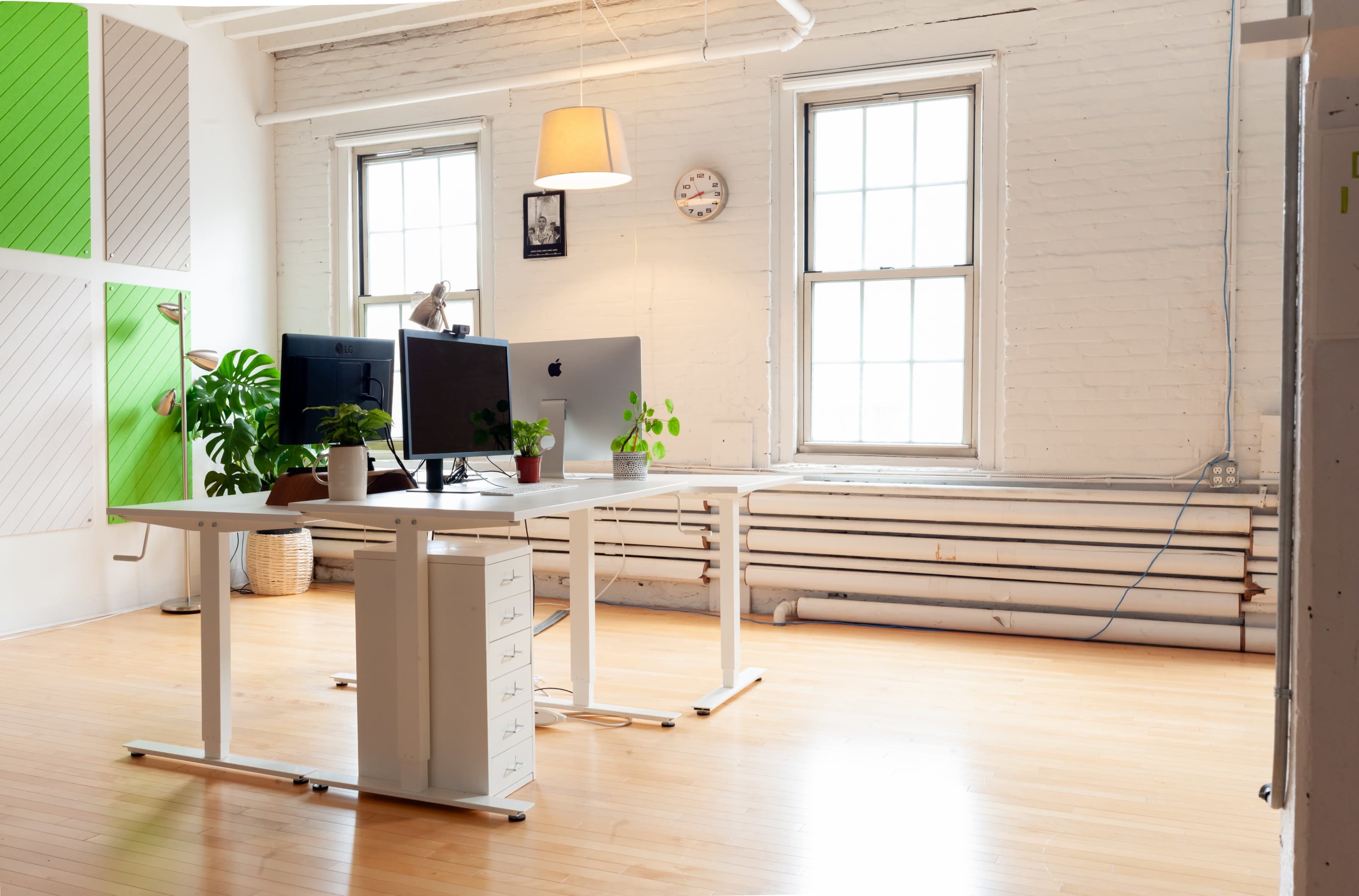 The image shows a modern office space with two desks, two monitors, a plant, and large windows allowing natural light to enter.