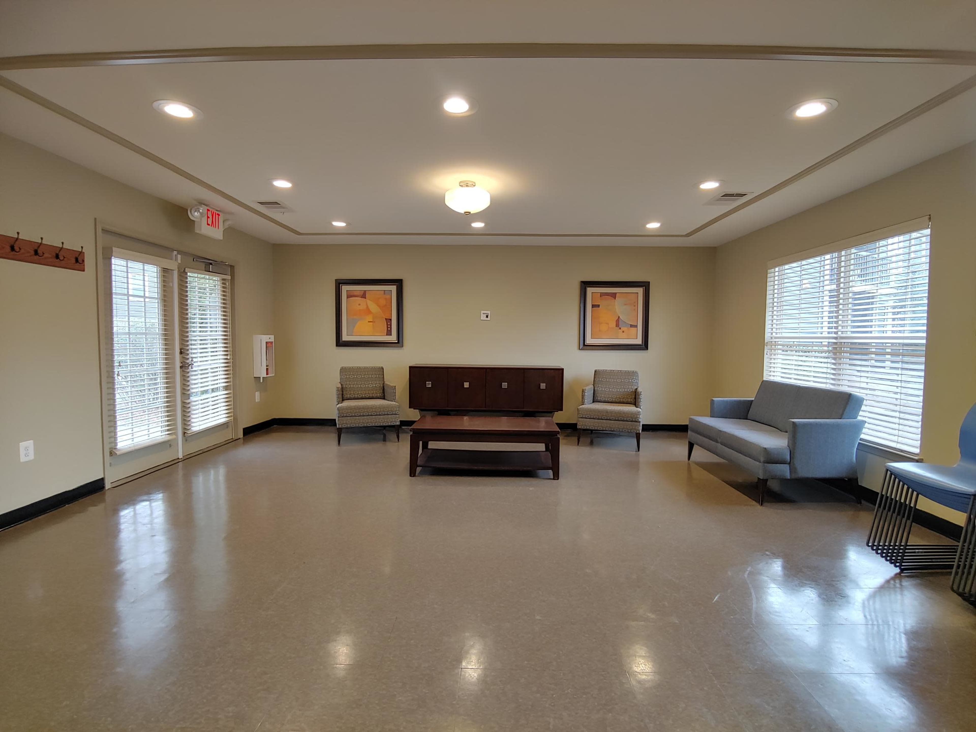 The image shows a spacious, well-lit room with two chairs, a sofa, a coffee table, and artwork on the walls, along with large windows allowing natural light in.