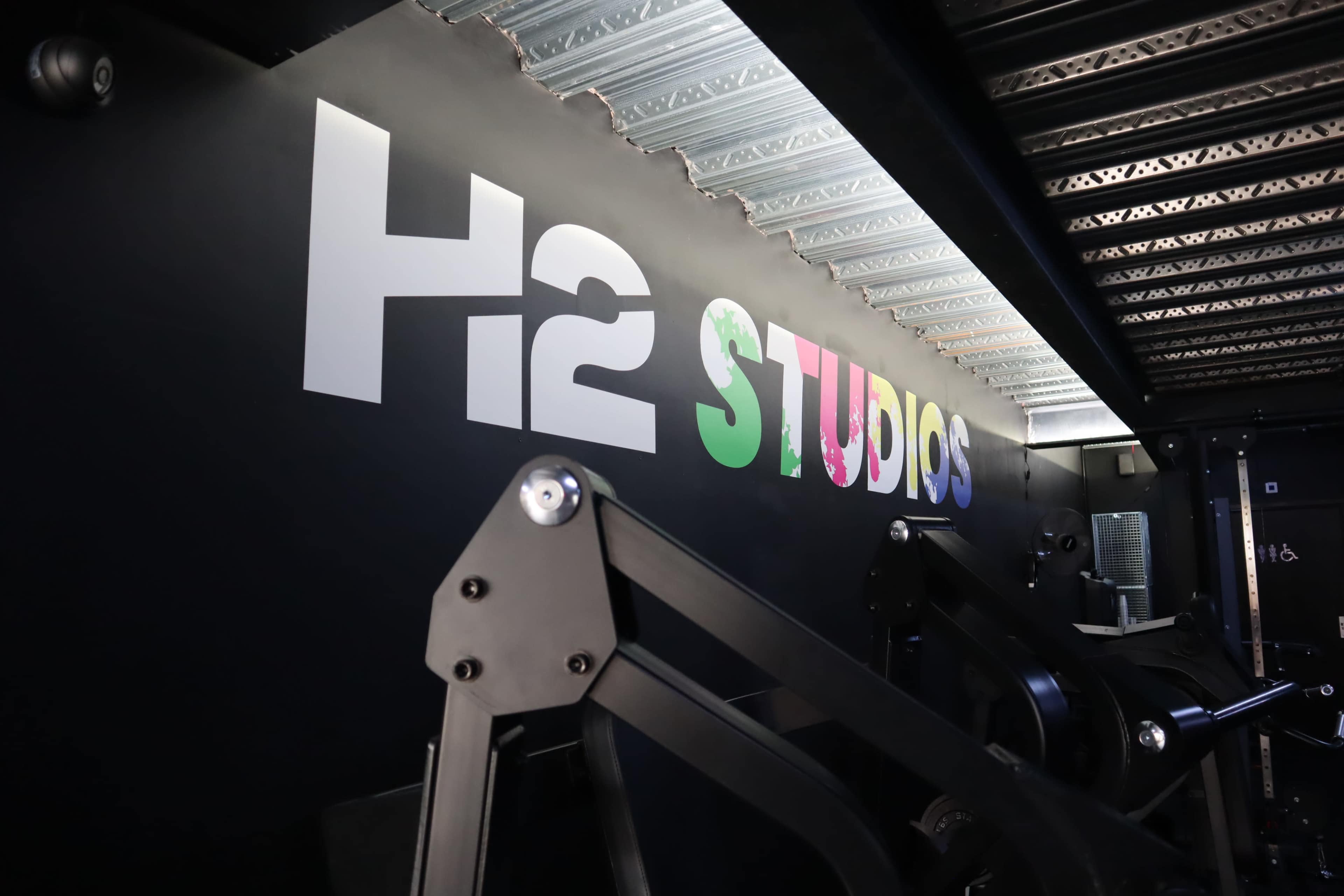 The image shows a black wall featuring the text "H2 STUDIOS" with colorful letters, alongside some exercise equipment.