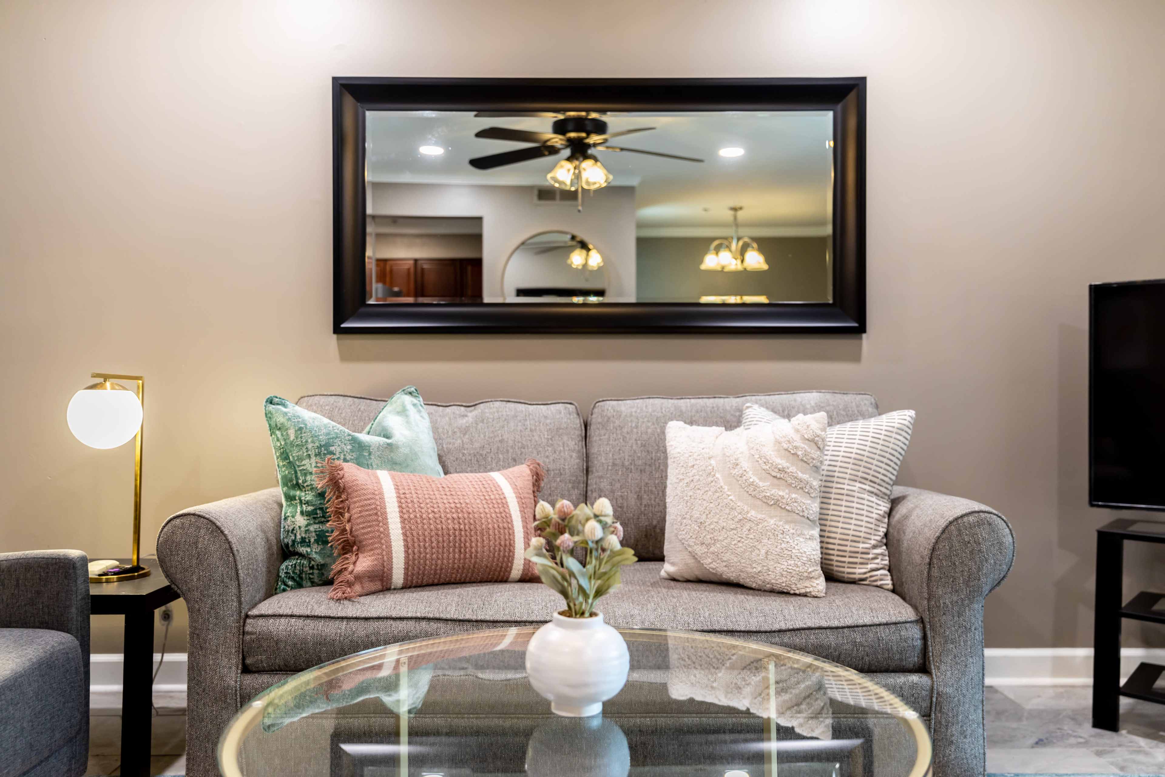 A gray sofa with various decorative pillows sits in front of a large mirror, reflecting light fixtures and a kitchen area in a living room.