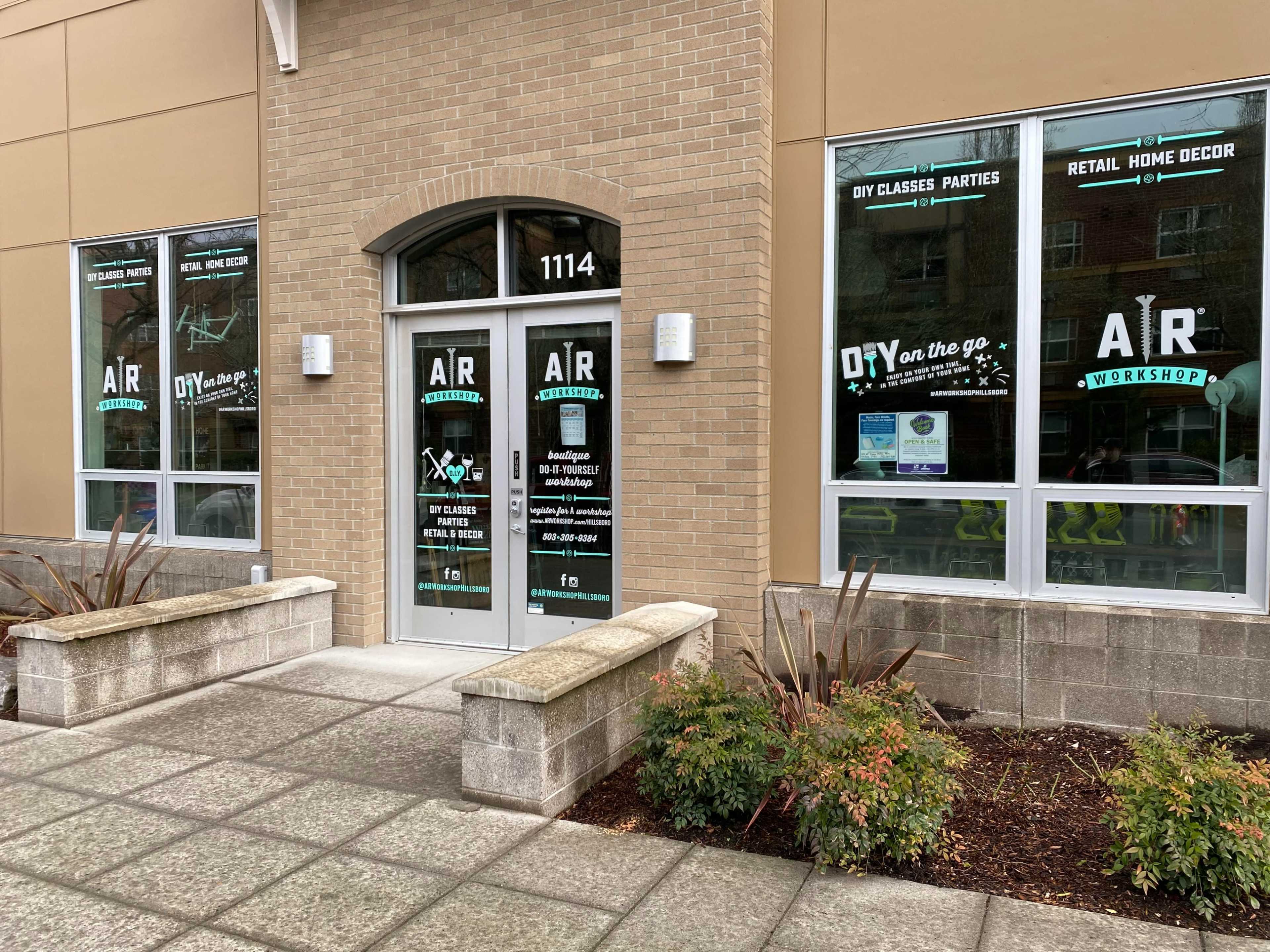 A storefront with large glass doors and windows displaying the business name "AR Workshop" along with promotional graphics for DIY classes and home decor.