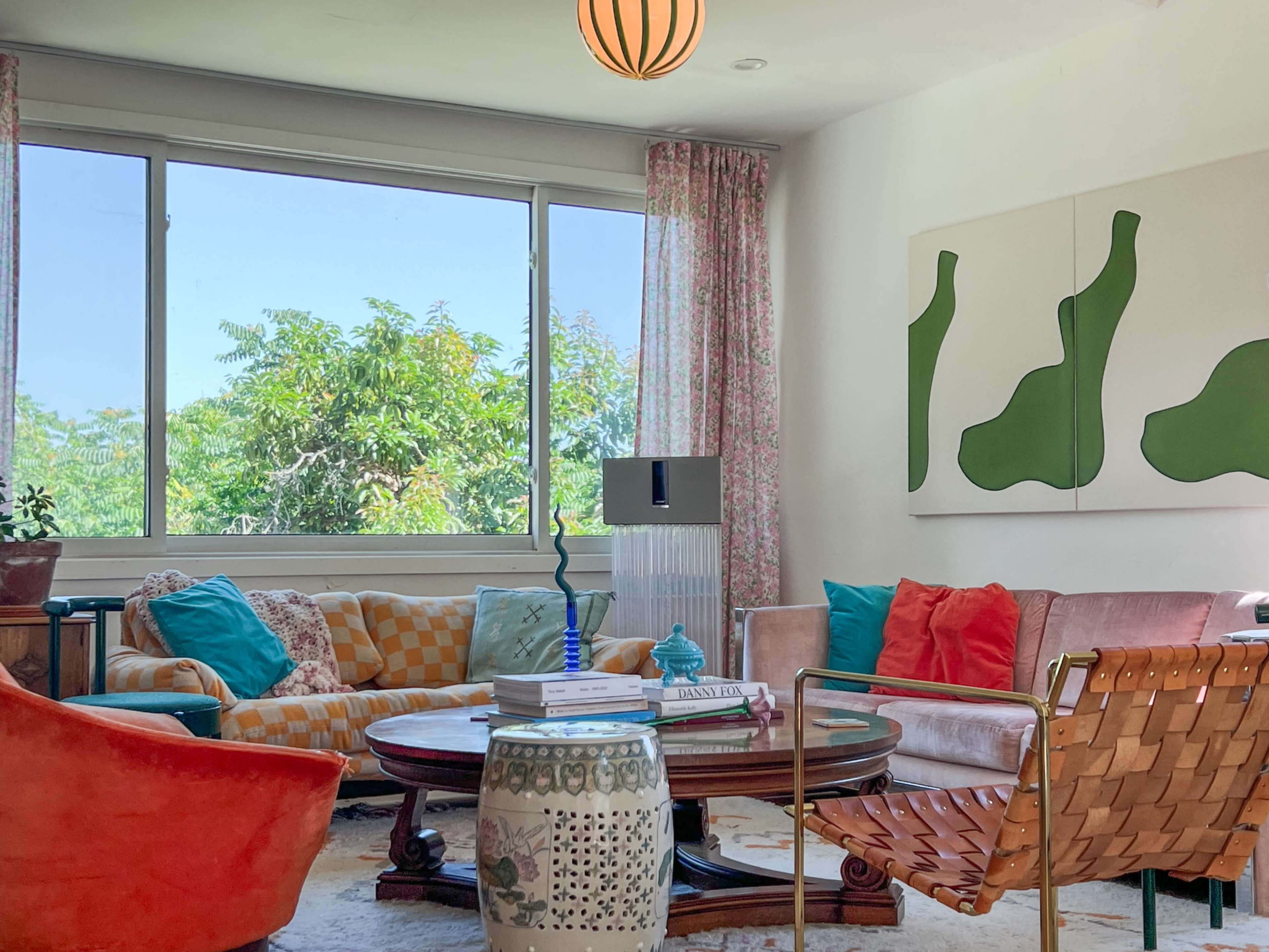 The living room features a mix of colorful furniture, including a patterned orange chair, a pink sofa, and a round coffee table, with large windows letting in natural light.