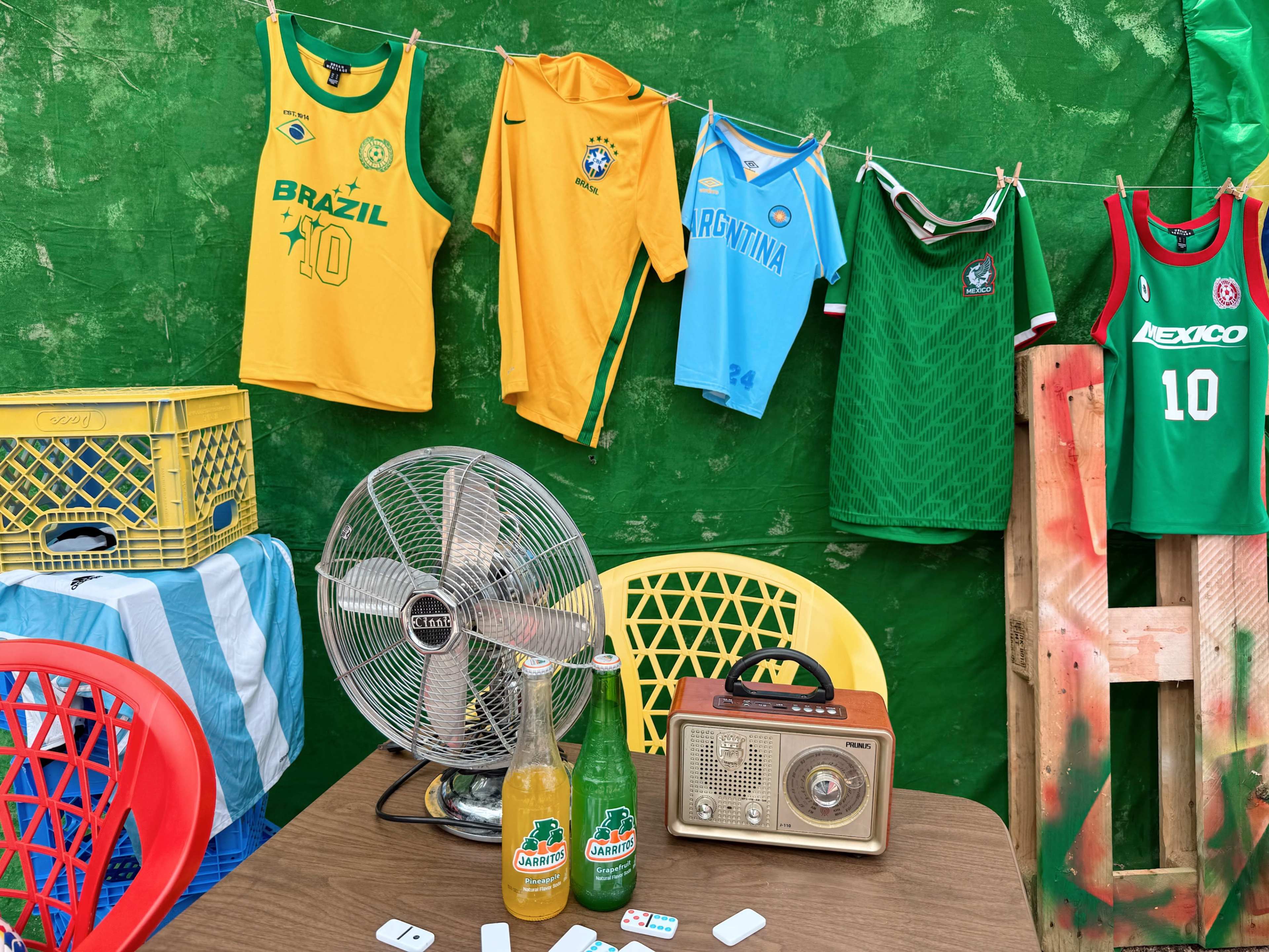 A colorful display of soccer jerseys hangs on a green wall, alongside a table featuring a vintage radio, two bottles, and a fan.
