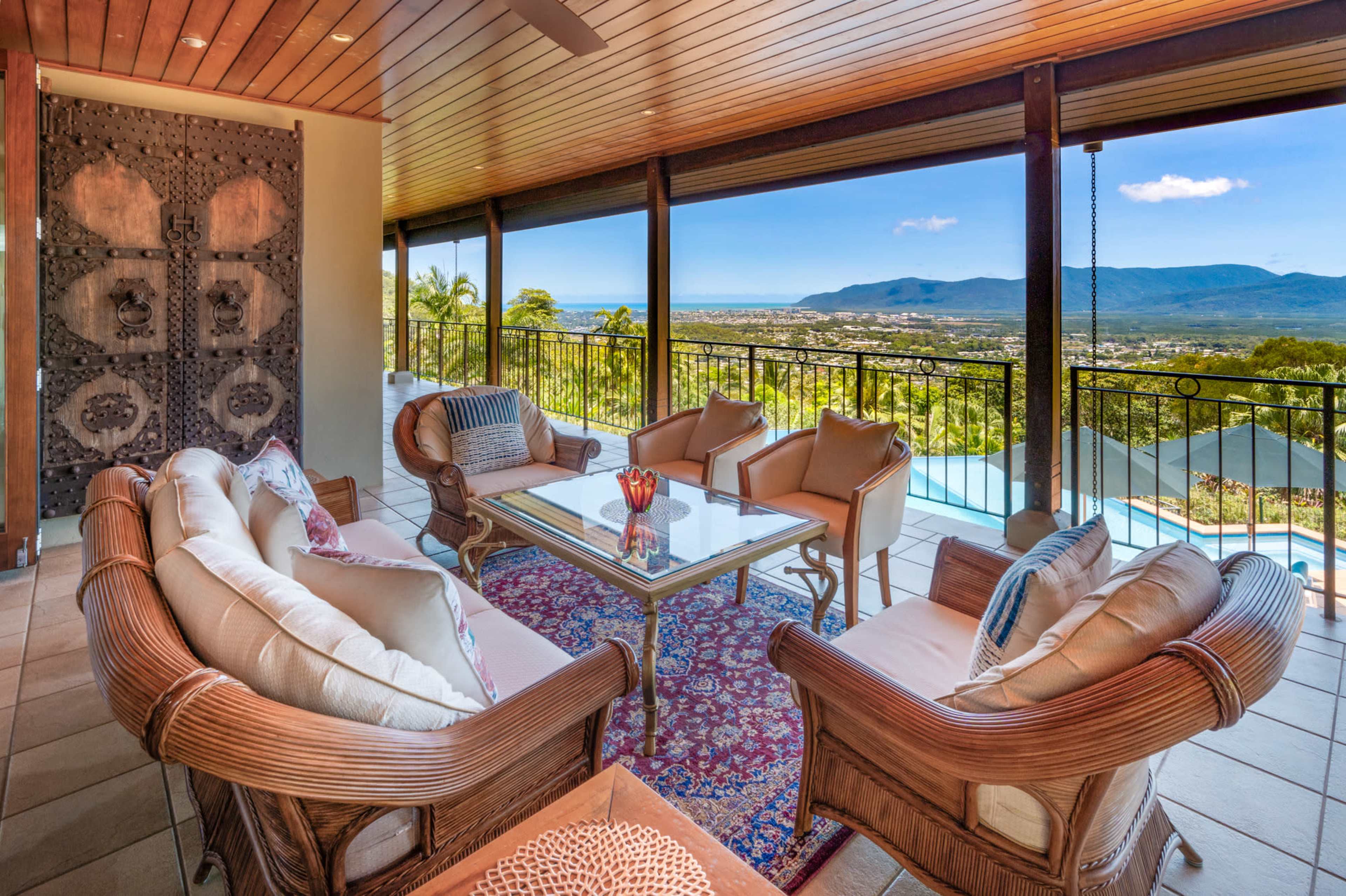 The image shows a spacious patio with a seating arrangement of wicker furniture, a glass table, and a scenic view of mountains and valleys.