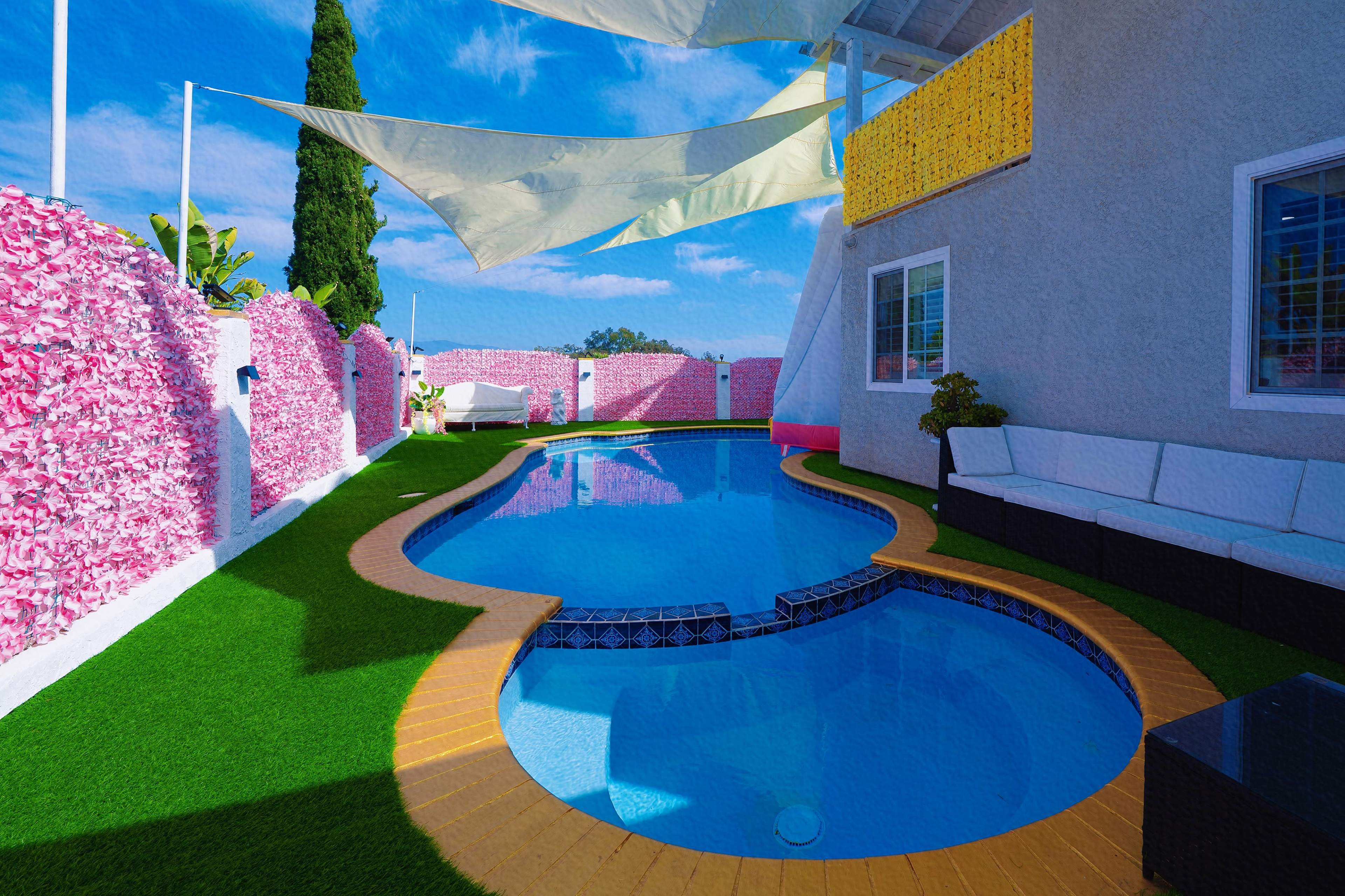 The image shows a vibrant backyard pool area with a blue swimming pool surrounded by artificial grass, pink flower-covered walls, and a modern seating arrangement.