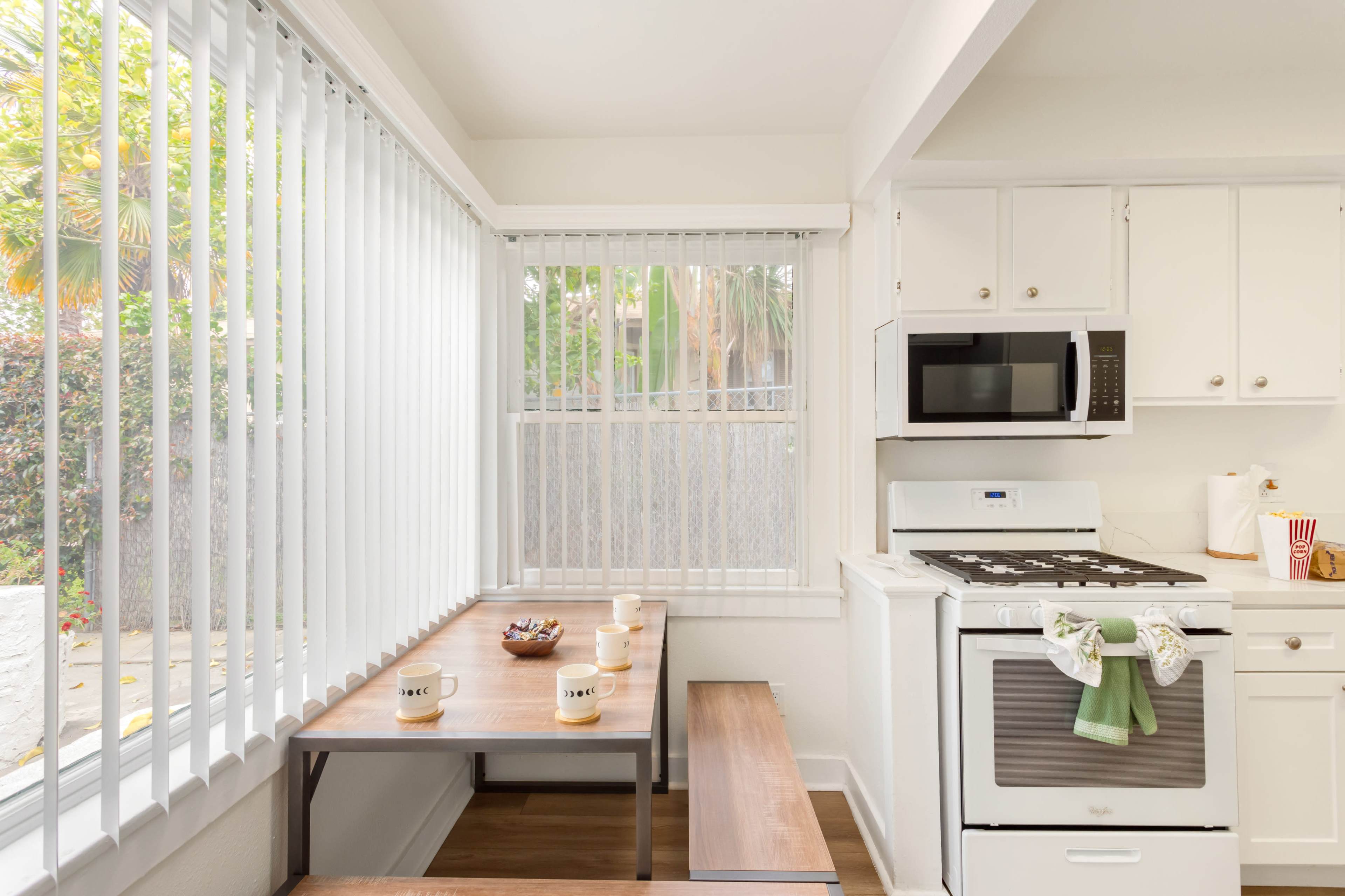 A small kitchen features a table with four mugs, a microwave, and a stove, illuminated by natural light through vertical blinds.