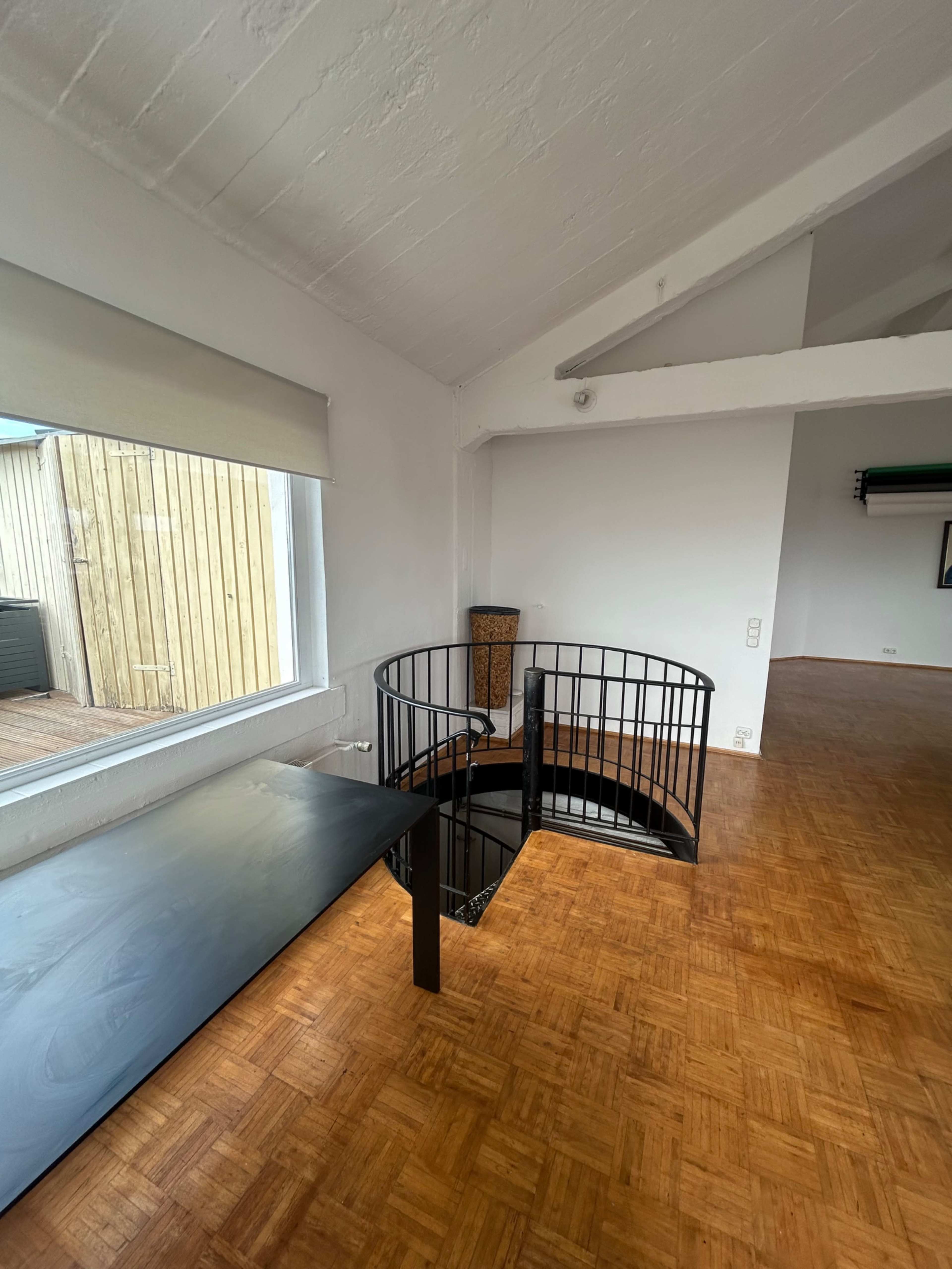 The image shows a spacious room with a circular staircase, a wooden floor, and large windows allowing natural light inside.