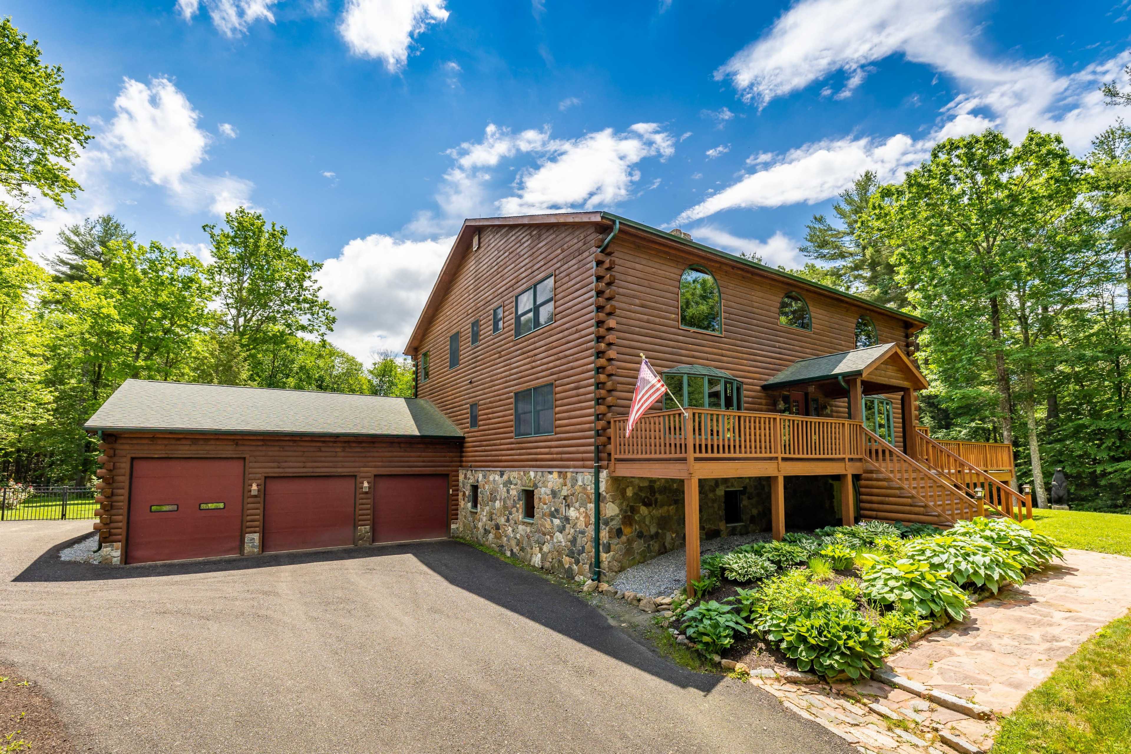 A large log cabin-style house features a stone and wood exterior, a front deck with stairs, and a two-car garage on a tree-lined property.