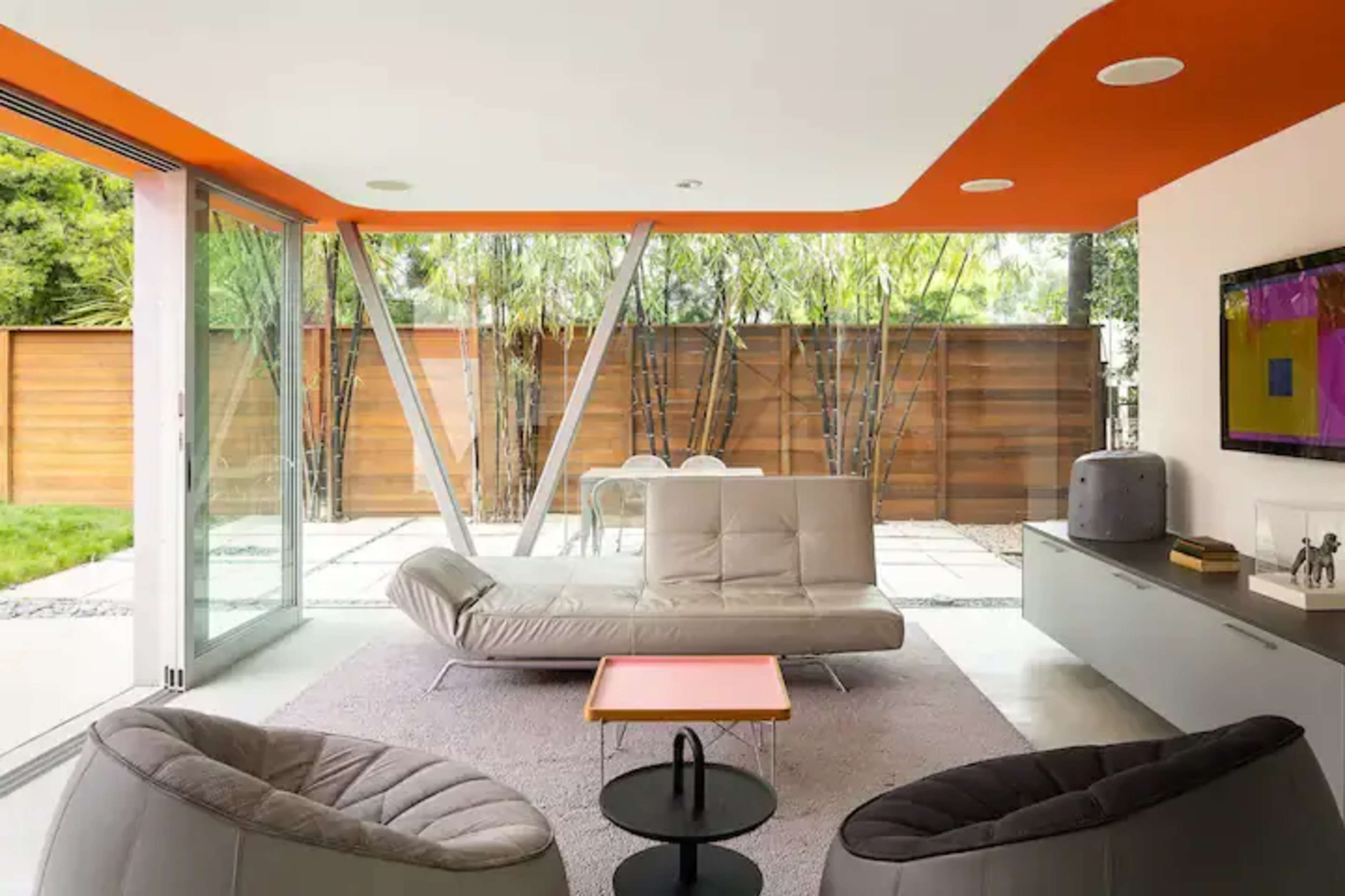 A modern living room features a sleek gray sofa, black armchairs, and large glass doors that open to a garden with bamboo plants.