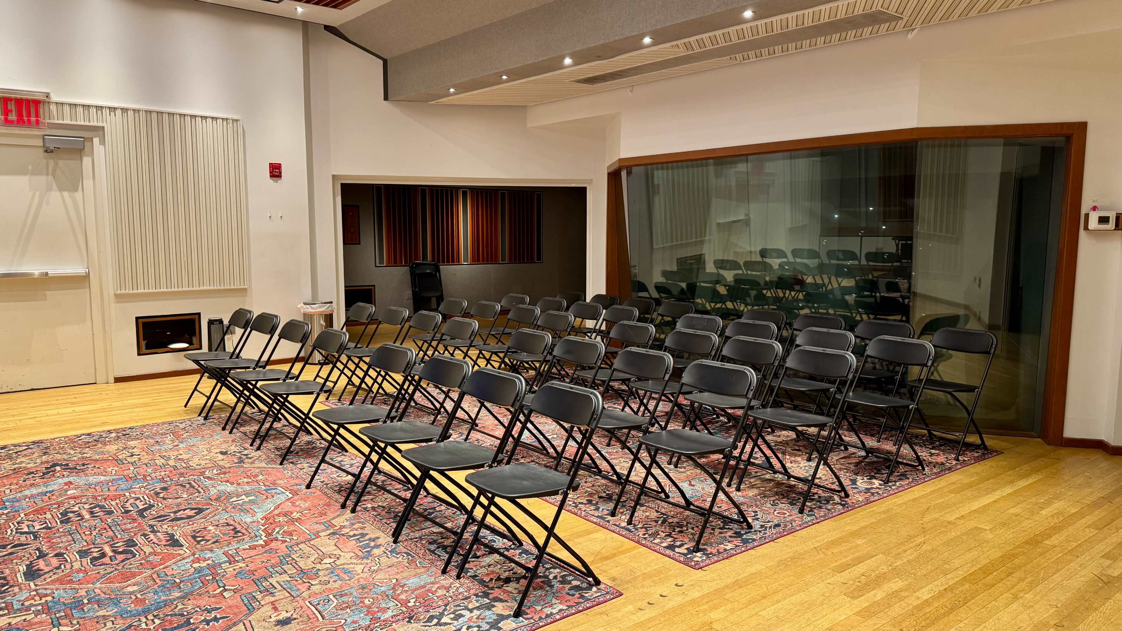 The image shows a room with a set of black folding chairs arranged in rows on a patterned rug, adjacent to a glass-walled space.