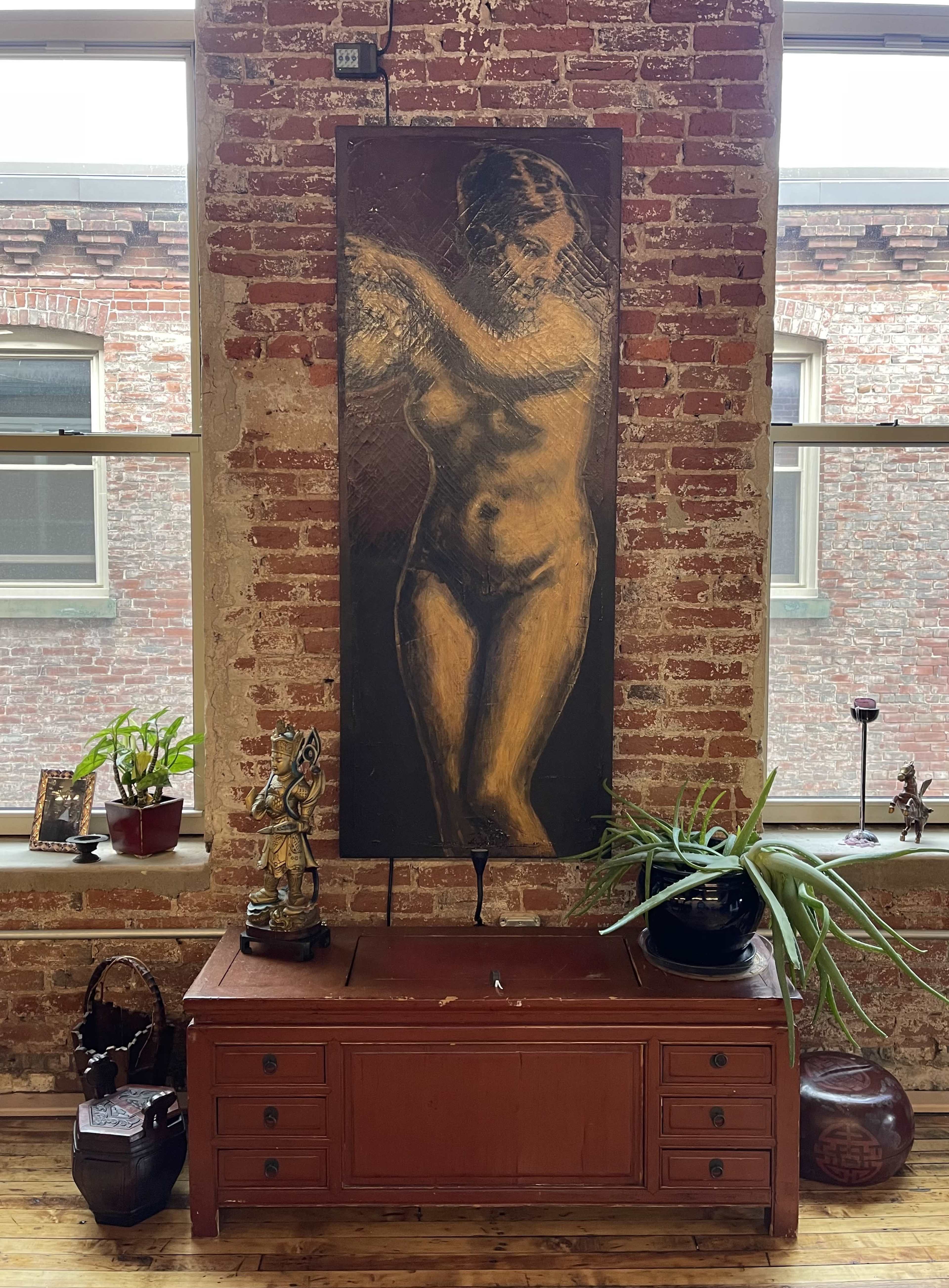 A large painting of a nude figure hangs on a brick wall above a wooden chest situated in a room filled with plants and decorative items.