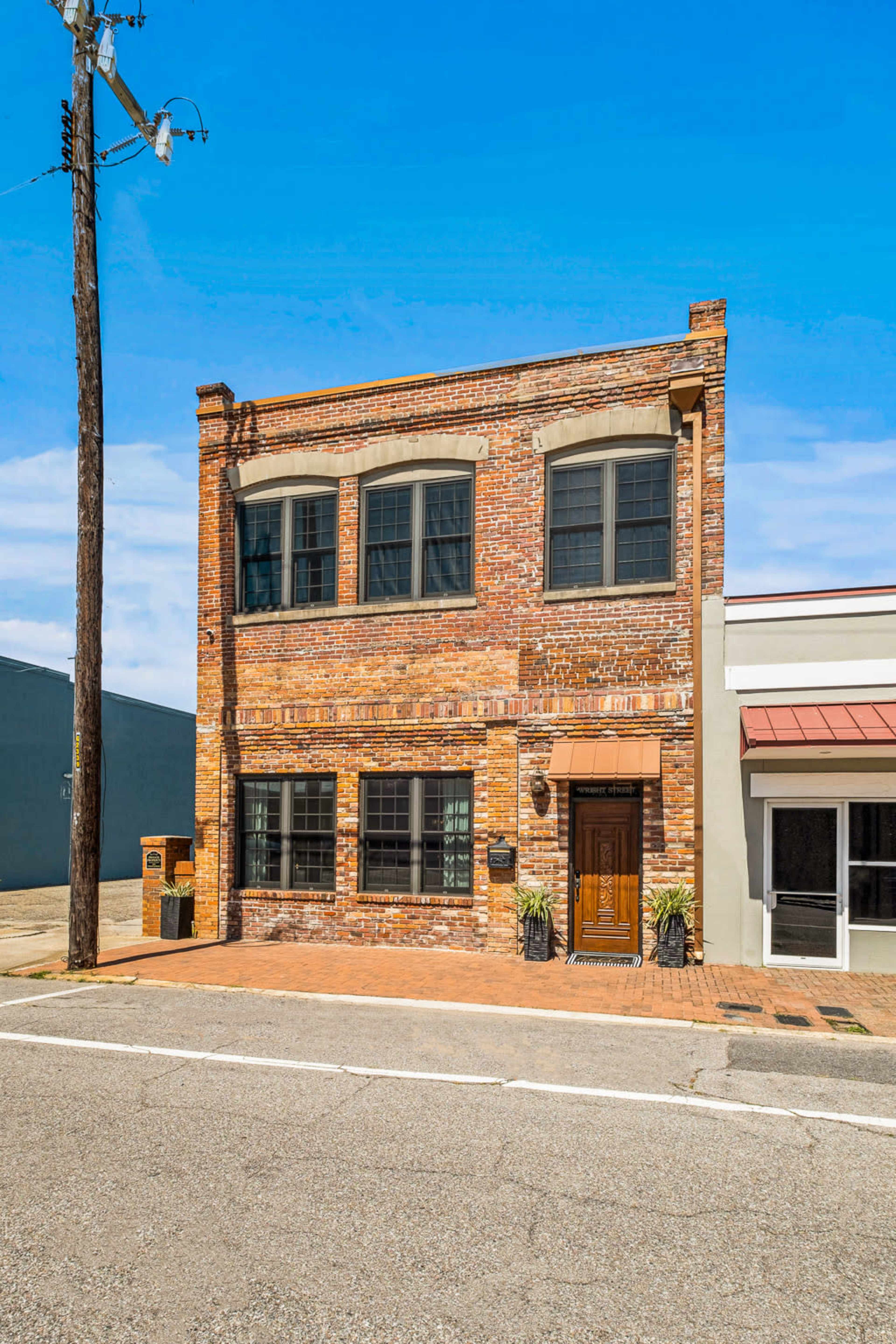 A two-story brick building with large windows and a wooden door stands on a street lined with a power pole.