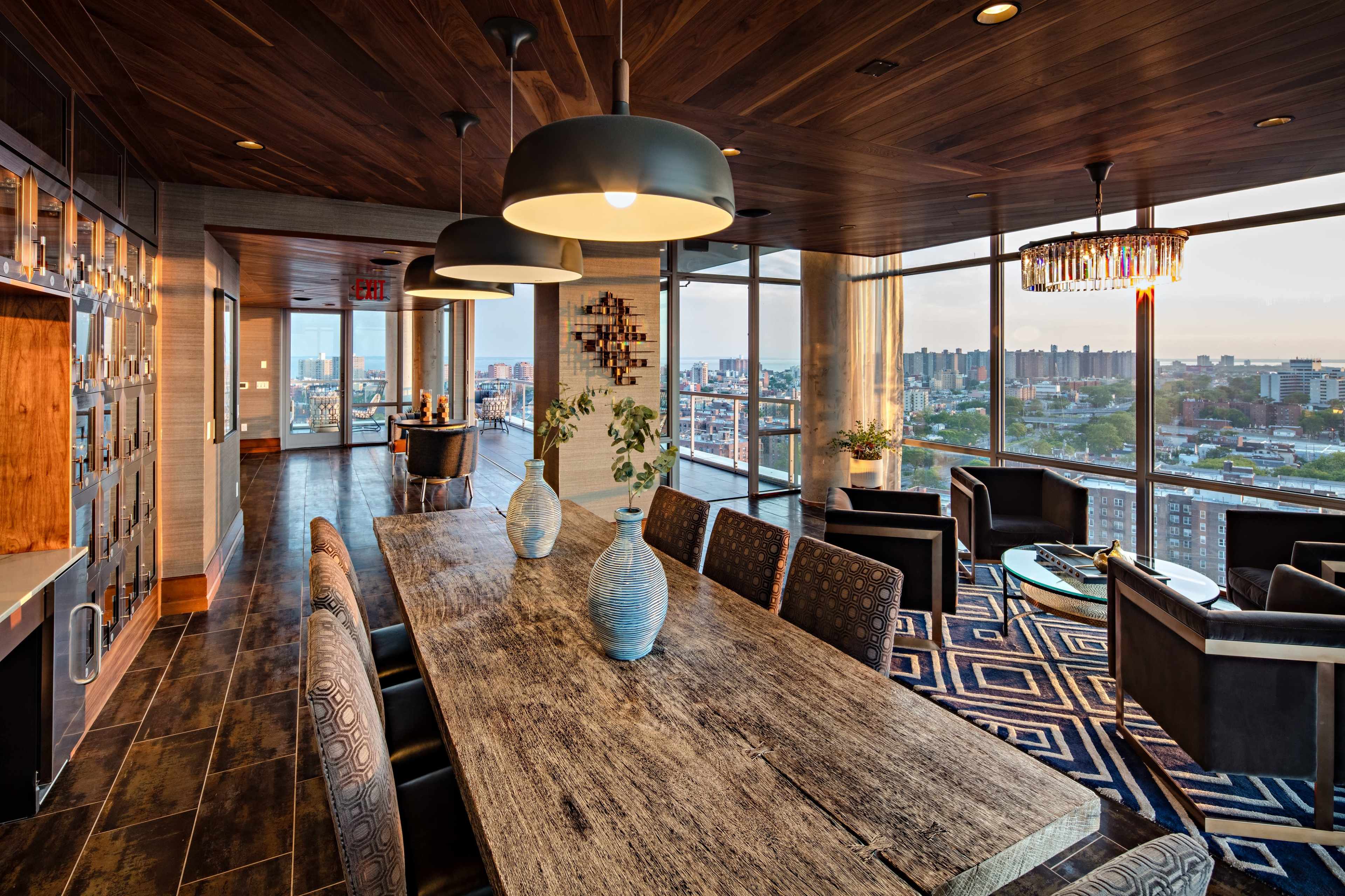 A modern dining area with a large wooden table, stylish chairs, and panoramic windows offering a view of the city skyline.
