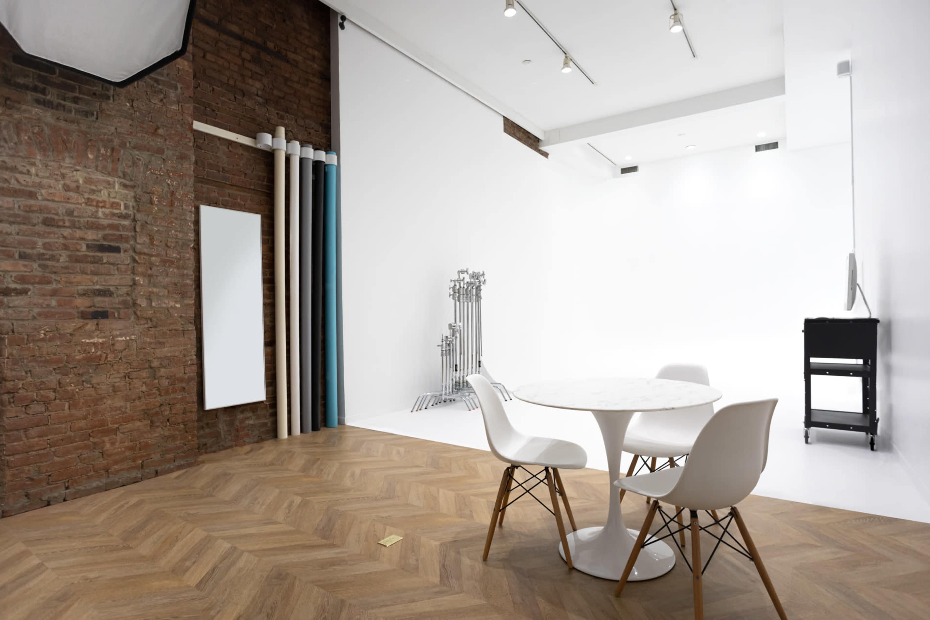 The image shows a modern photography studio with a round table and white chairs, featuring a brick wall and a large white backdrop.