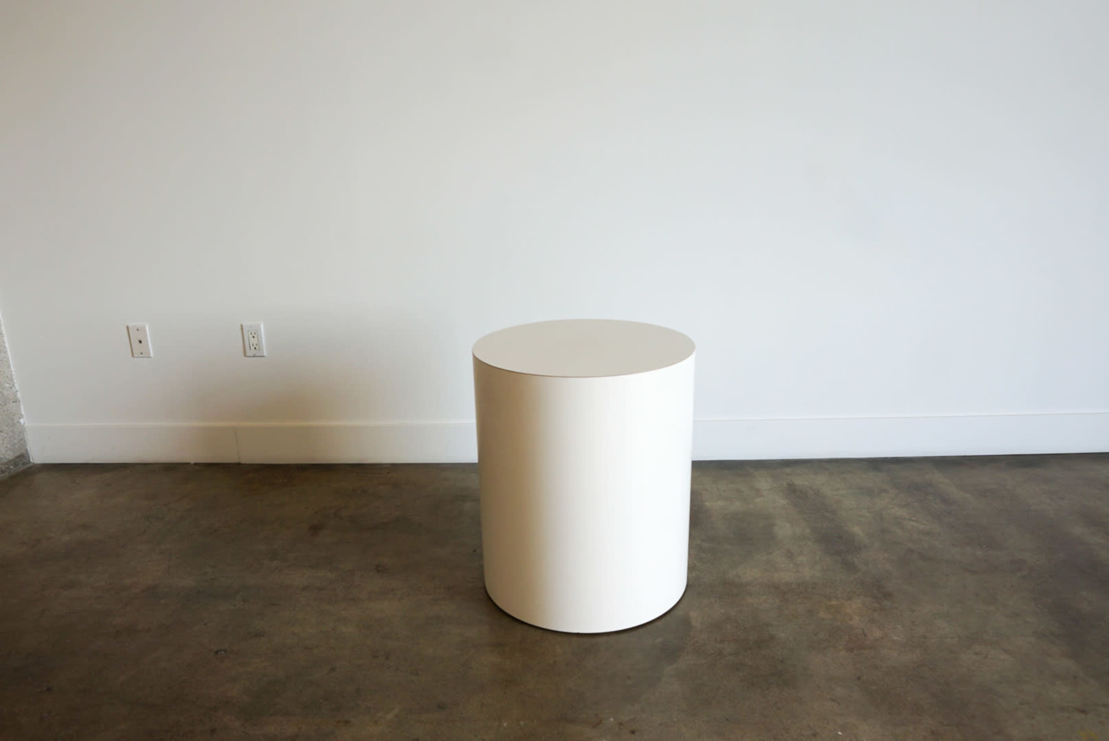 A cylindrical white table is positioned in front of a plain wall on a concrete floor.