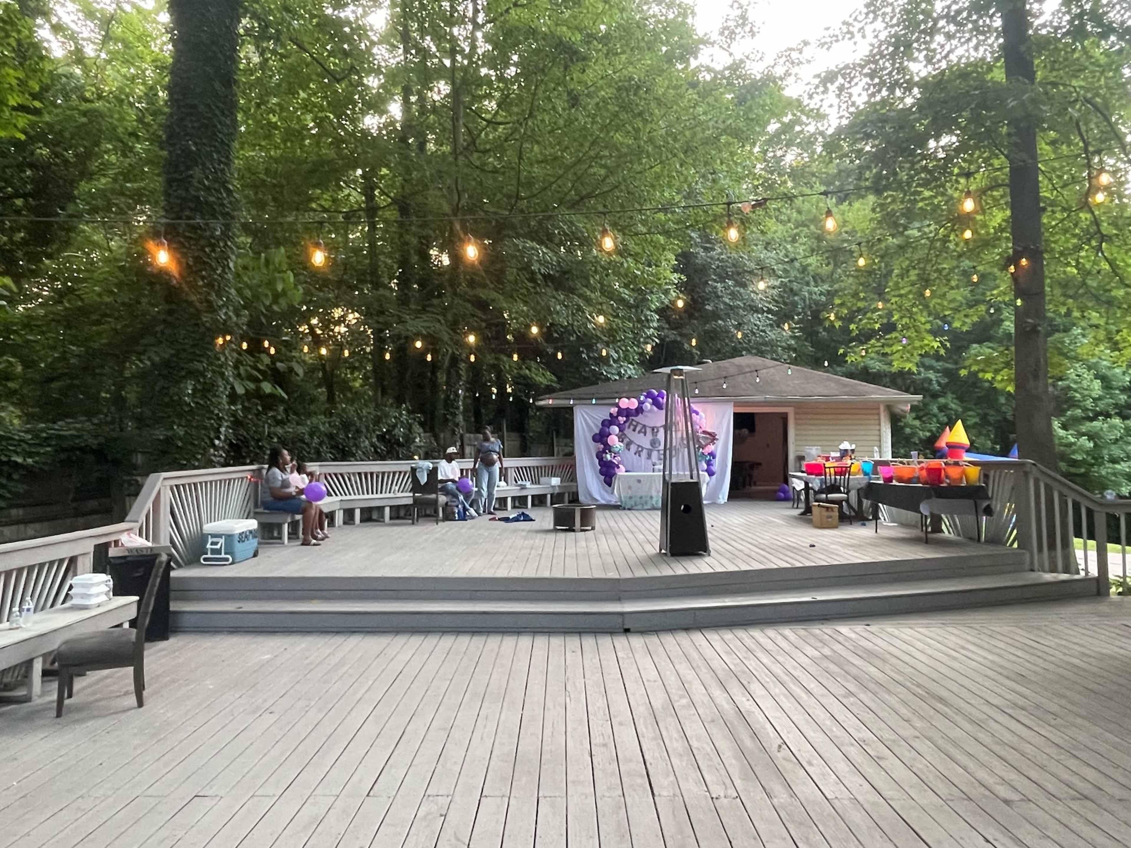 A wooden deck in a wooded area features string lights overhead, with people gathered near a decorated area and tables set up for an event.