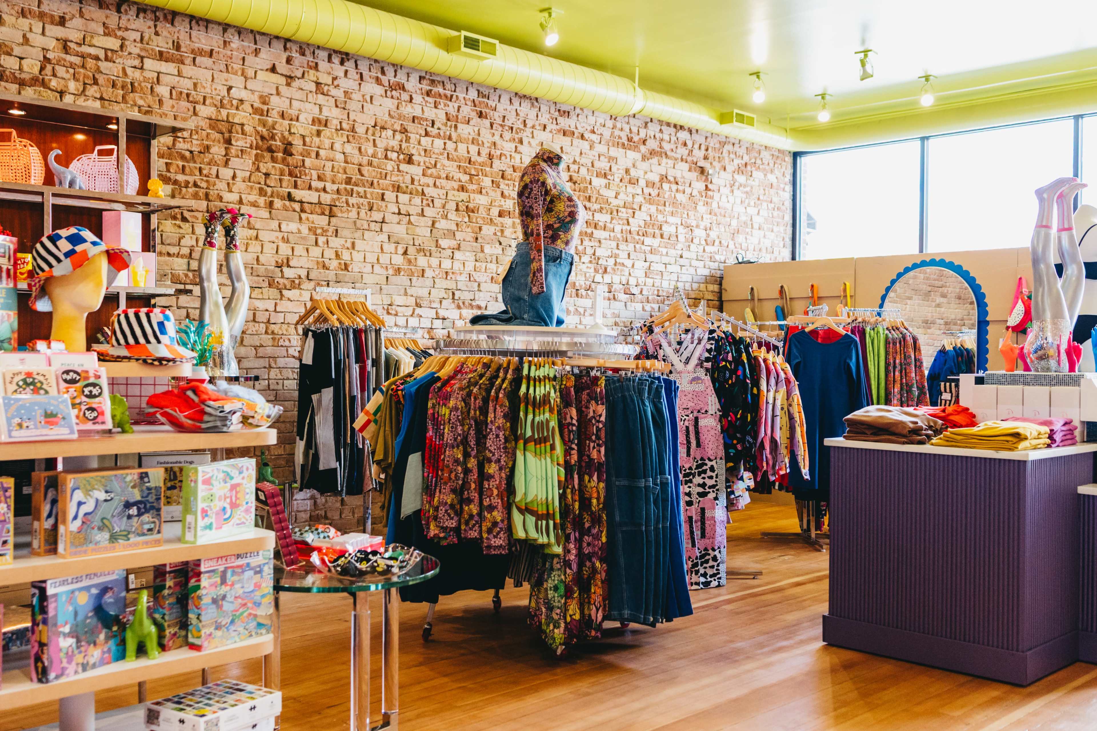 The image depicts a colorful boutique interior featuring a variety of clothing racks, accessories, and display shelves filled with items for sale, set against a brick wall.