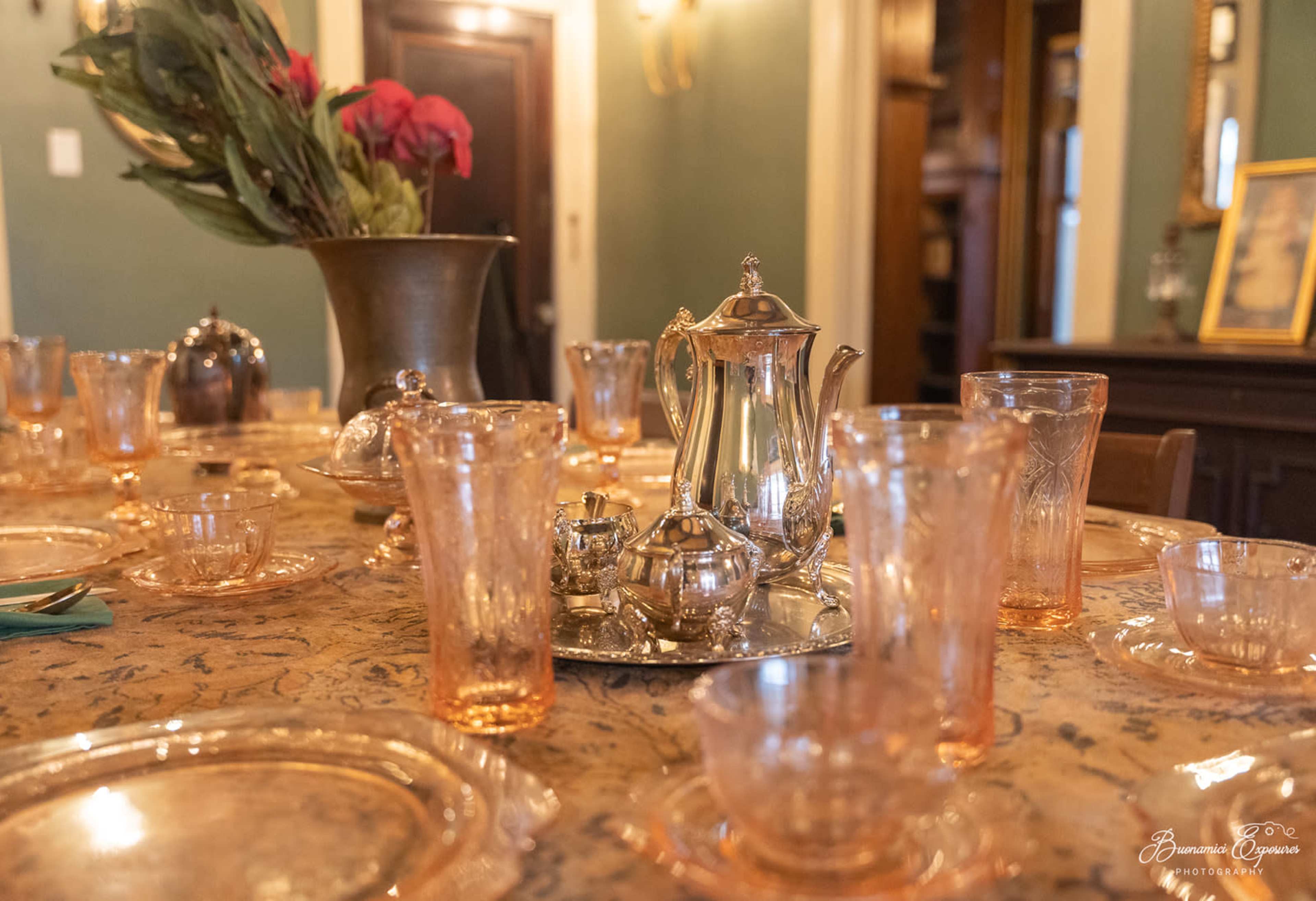 A polished silver tea set and delicate glassware are arranged on a table adorned with an ornate tablecloth and decorative floral arrangements.