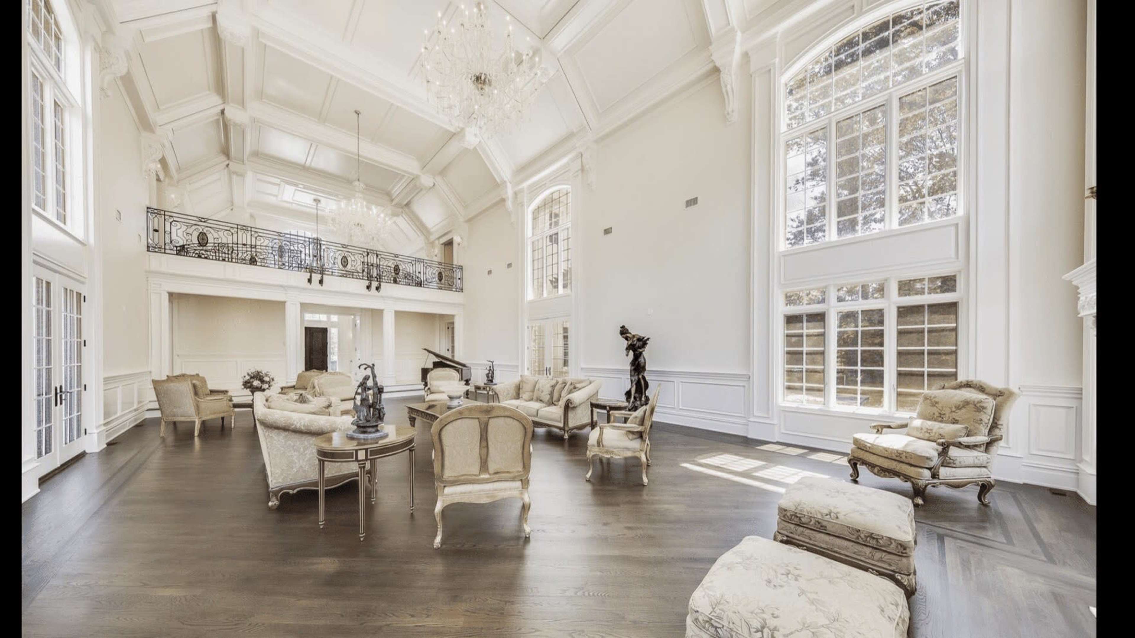 The image shows a spacious, elegantly furnished living room with high ceilings, large windows, and a grand chandelier.