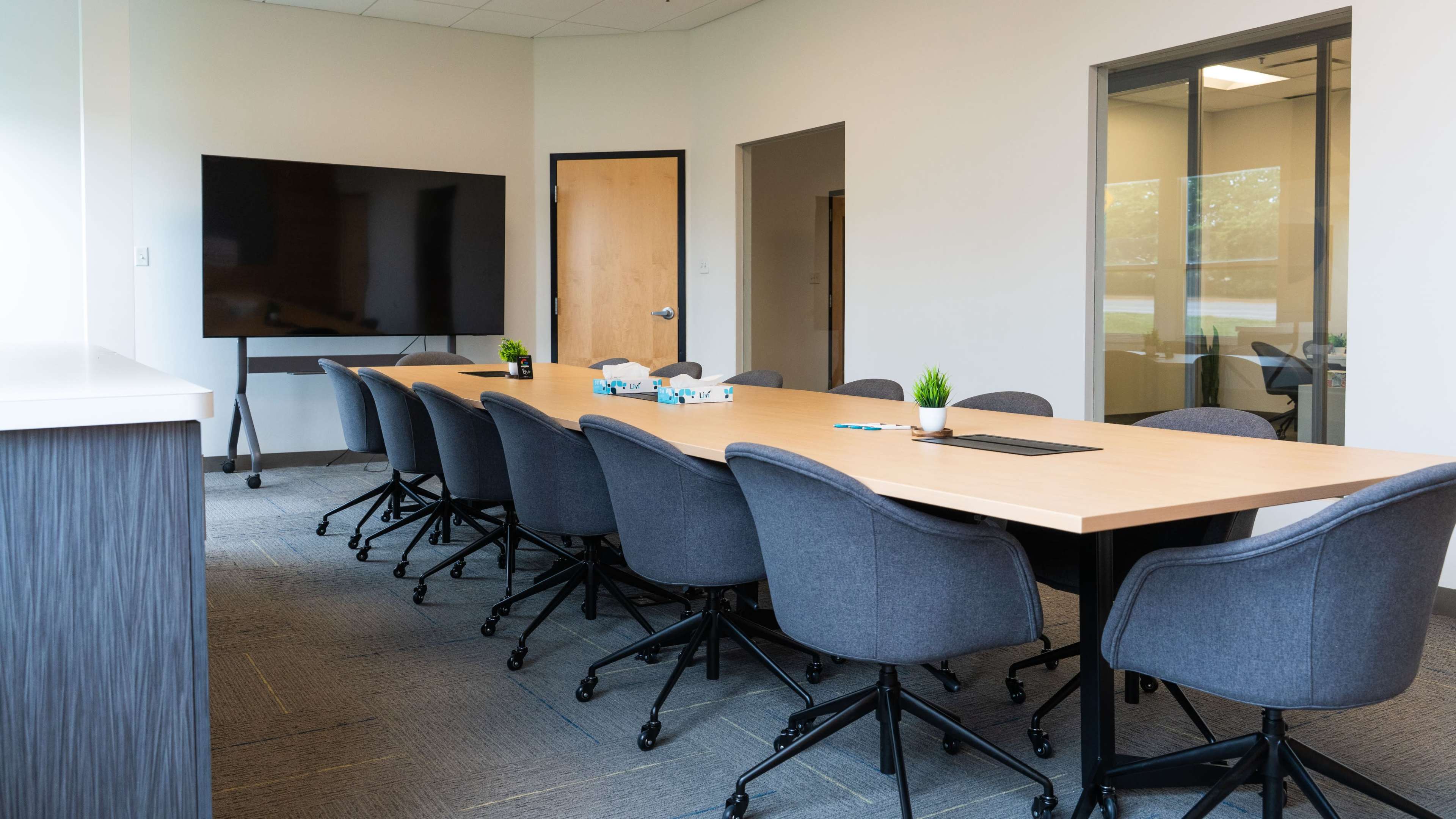 A large conference room features a long wooden table surrounded by gray rolling chairs, with a mounted television on the wall and plants on the table.