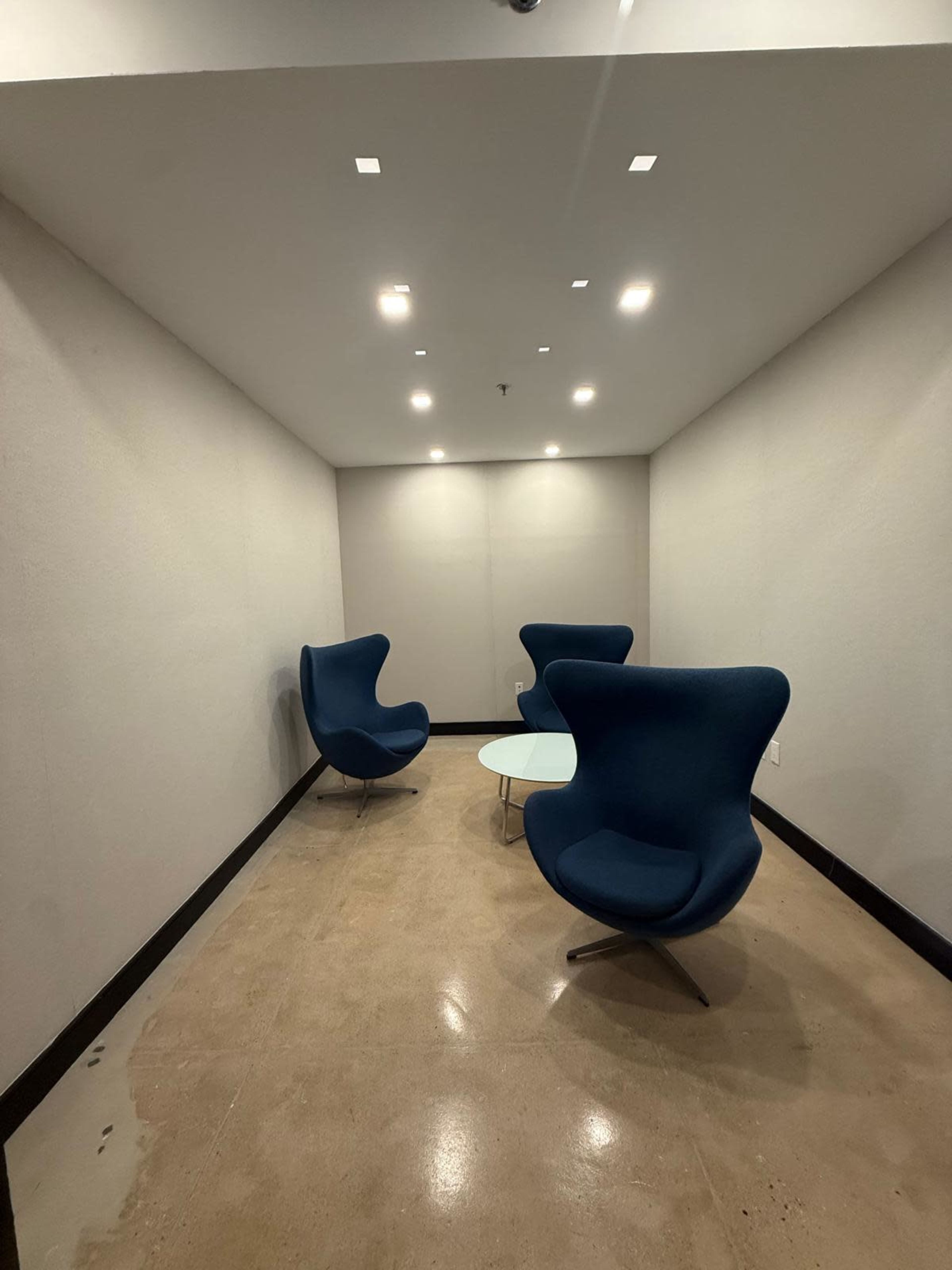 The image shows a small, minimalist meeting room featuring two blue chairs and a glass table under recessed lighting.