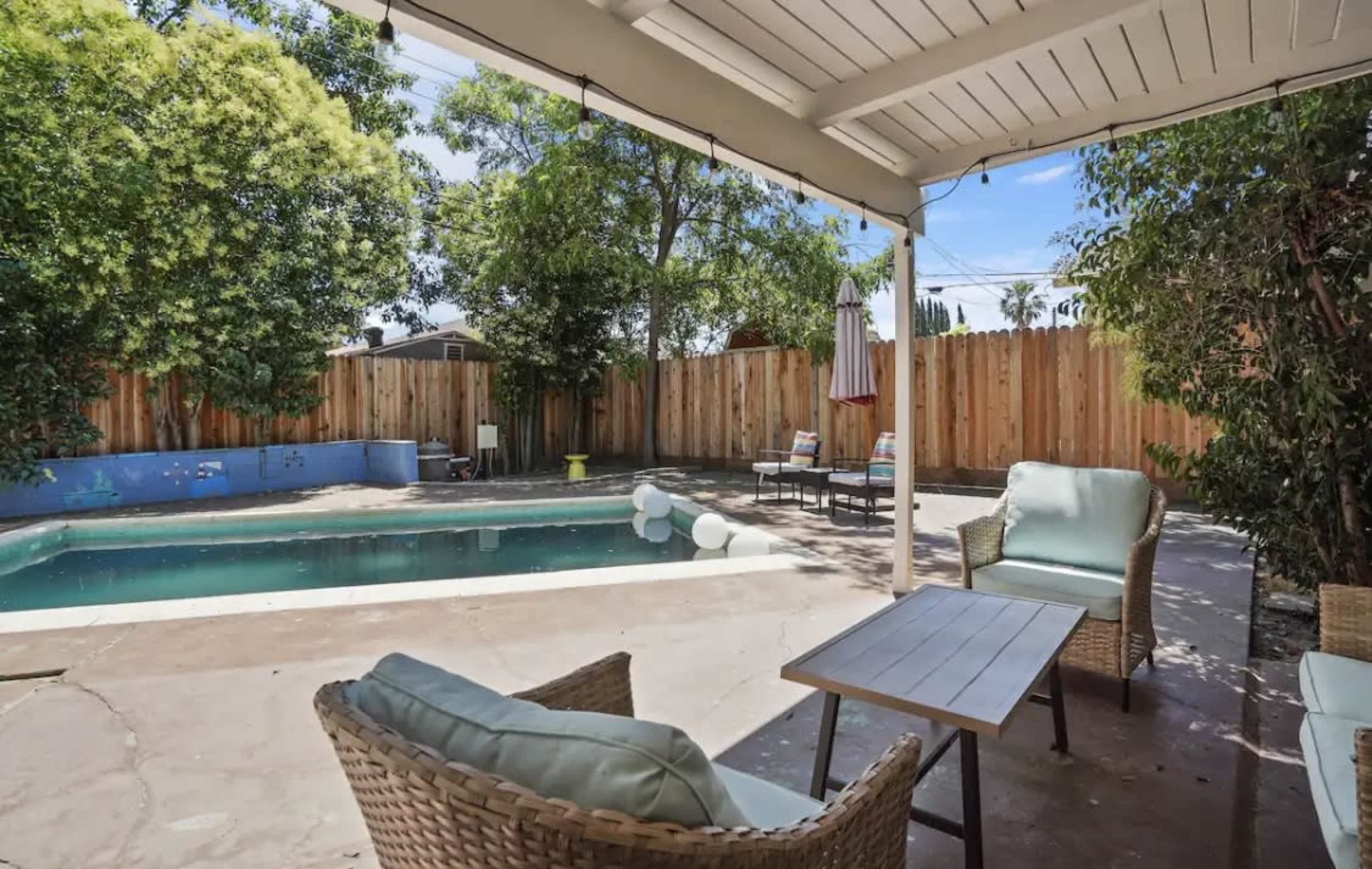 A backyard with a swimming pool, surrounded by a wooden fence, and furnished with a pair of wicker chairs and a small table.