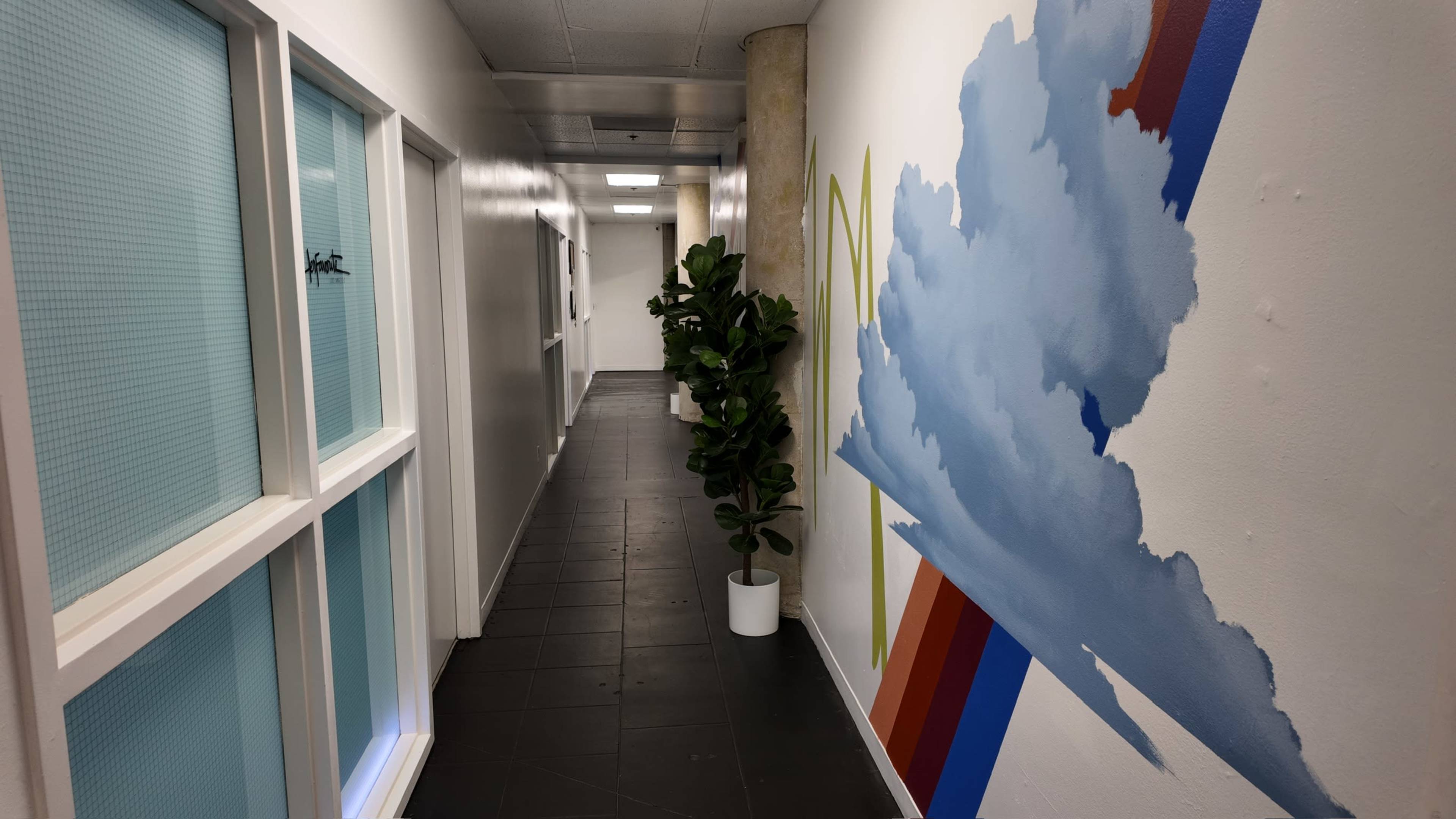 A corridor features a mural of clouds on one wall, with doors leading to adjacent rooms and a potted plant positioned along the side.