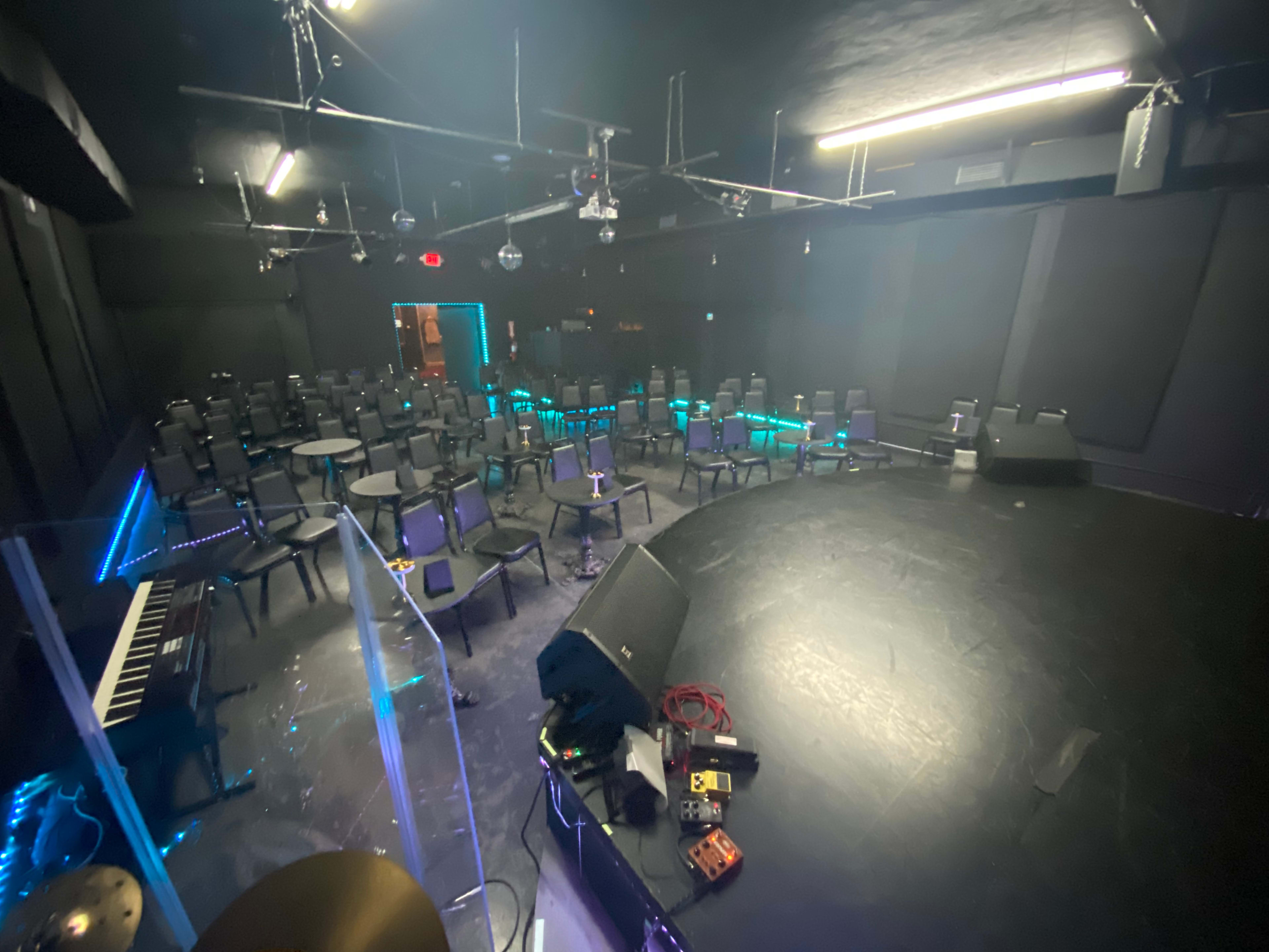 The image shows a dimly lit performance space with rows of empty chairs facing a small stage, featuring a keyboard and various music equipment scattered on the floor.