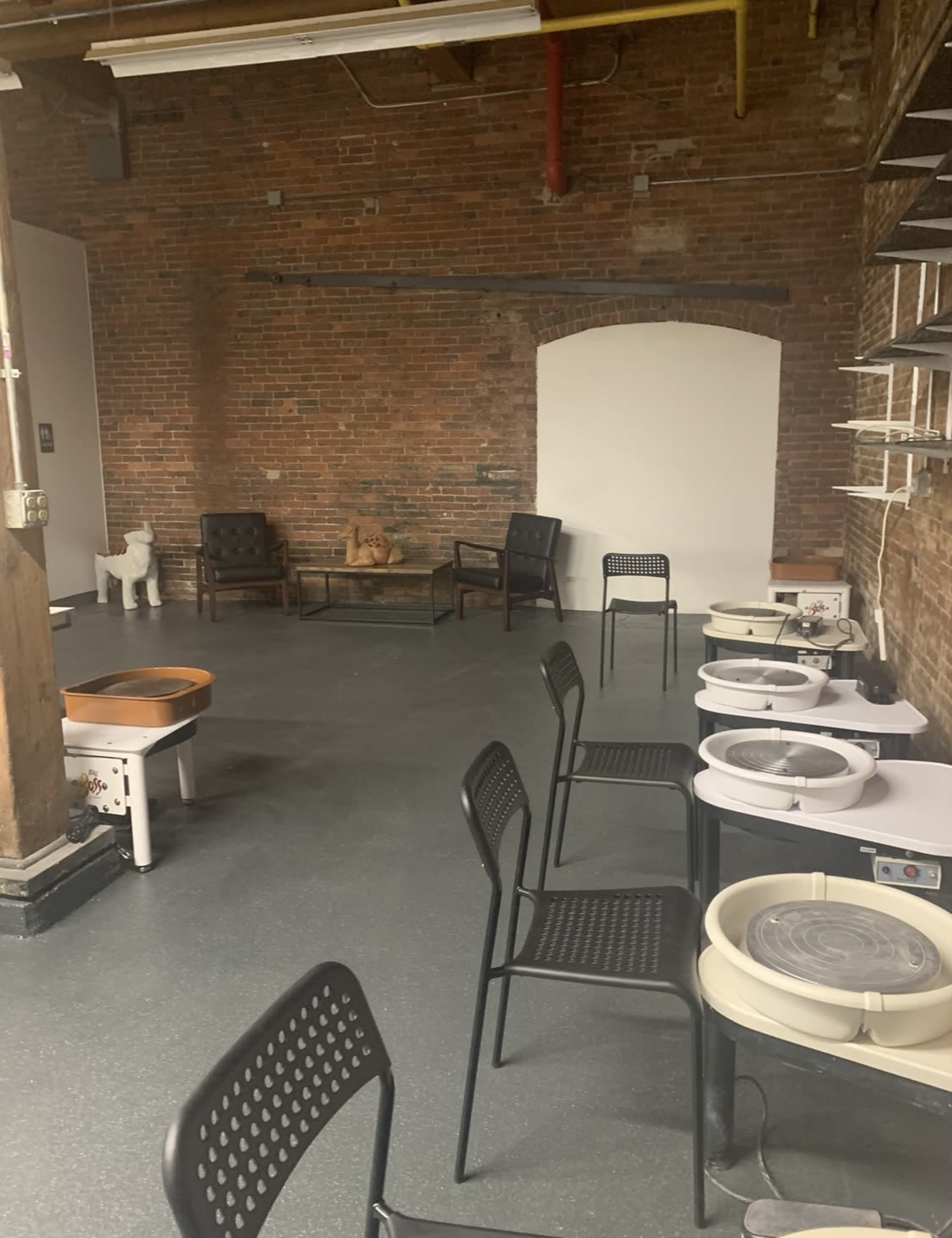 An interior space with exposed brick walls, several pottery wheels, and a few chairs arranged around a wooden table.