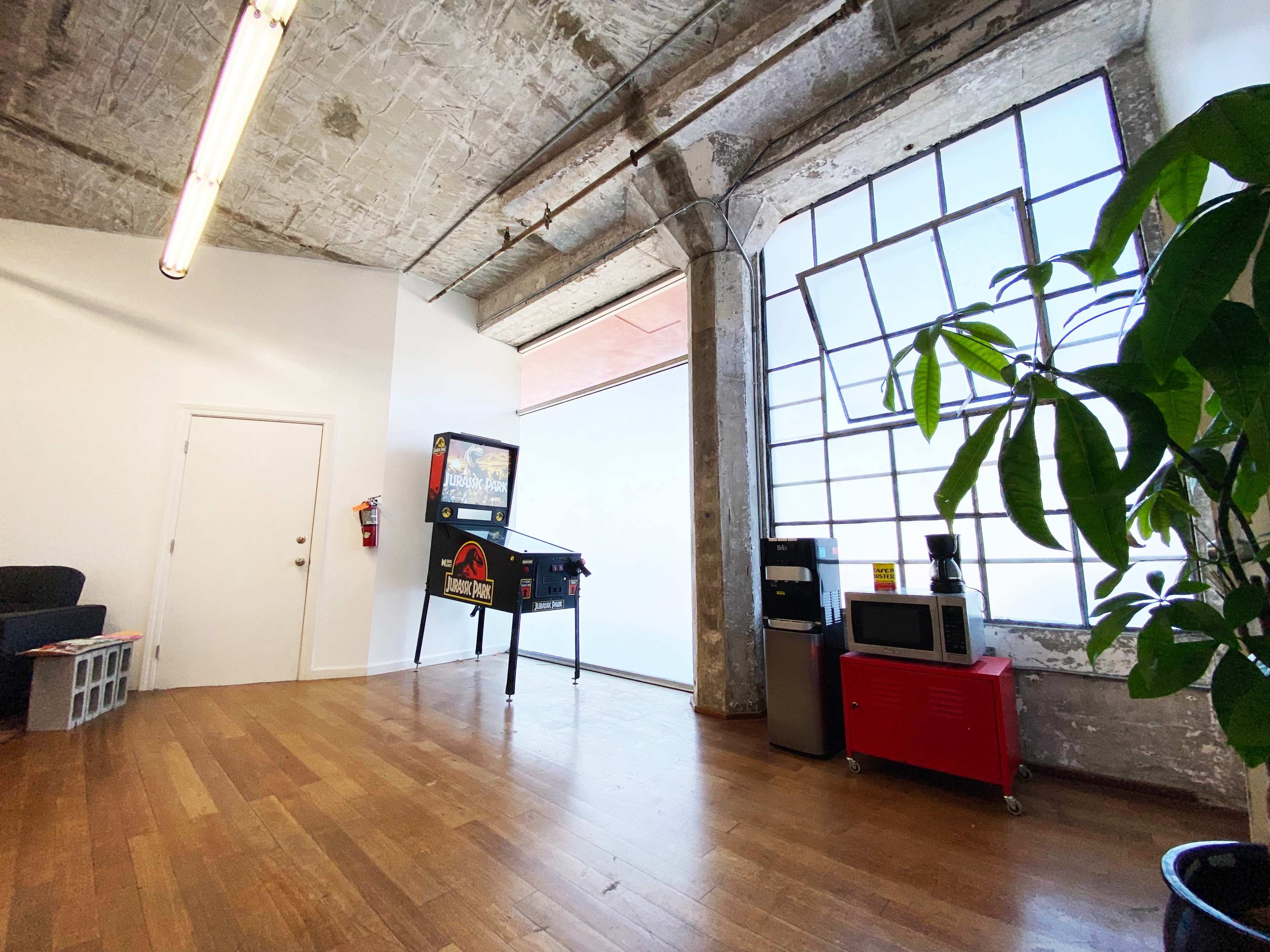 An open room with a pinball machine, a microwave, a red cart, and a large window alongside a potted plant.