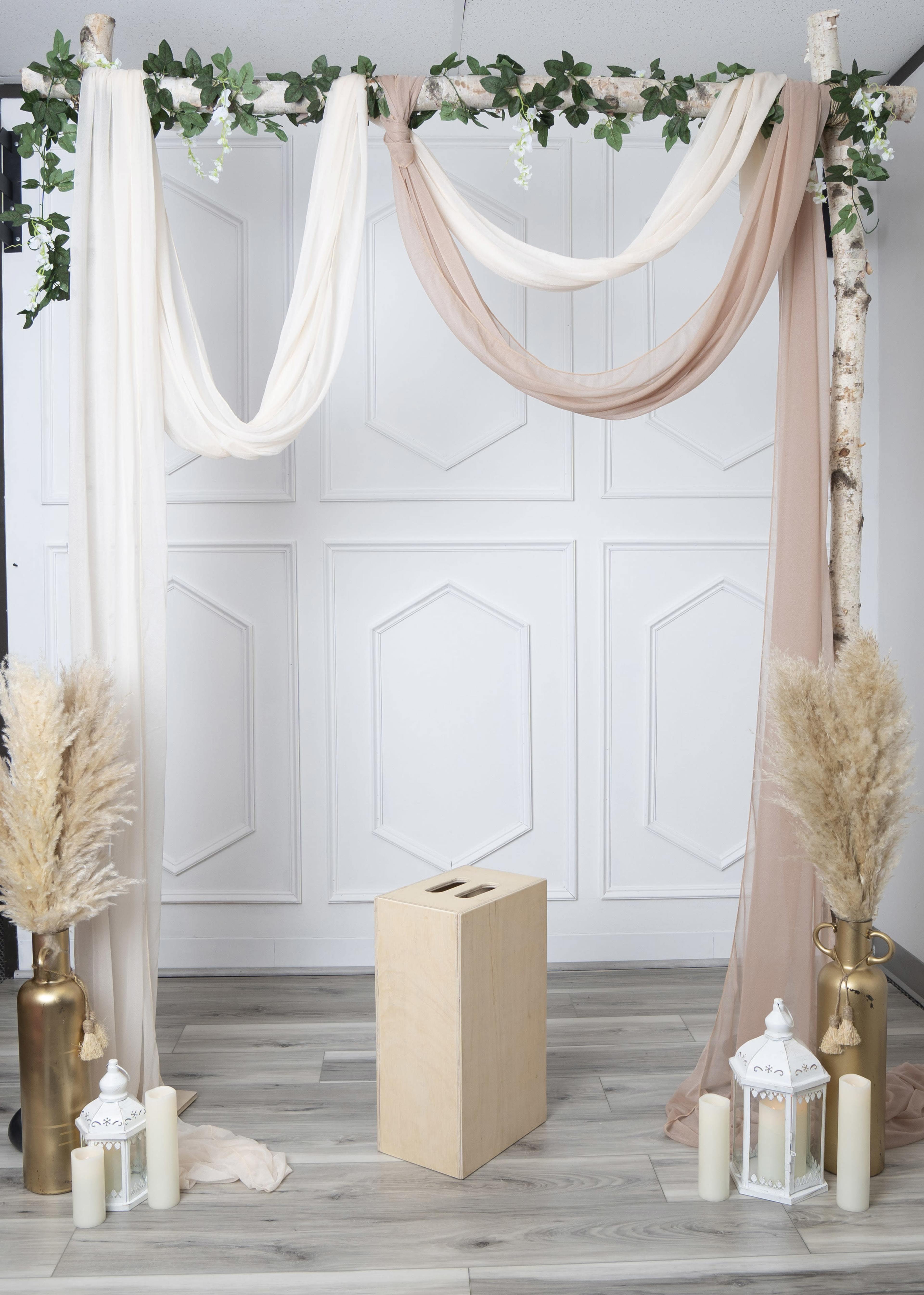 The image shows a decorated backdrop with draped fabrics, a wooden pedestal, and decorative elements including lanterns and dried plants.