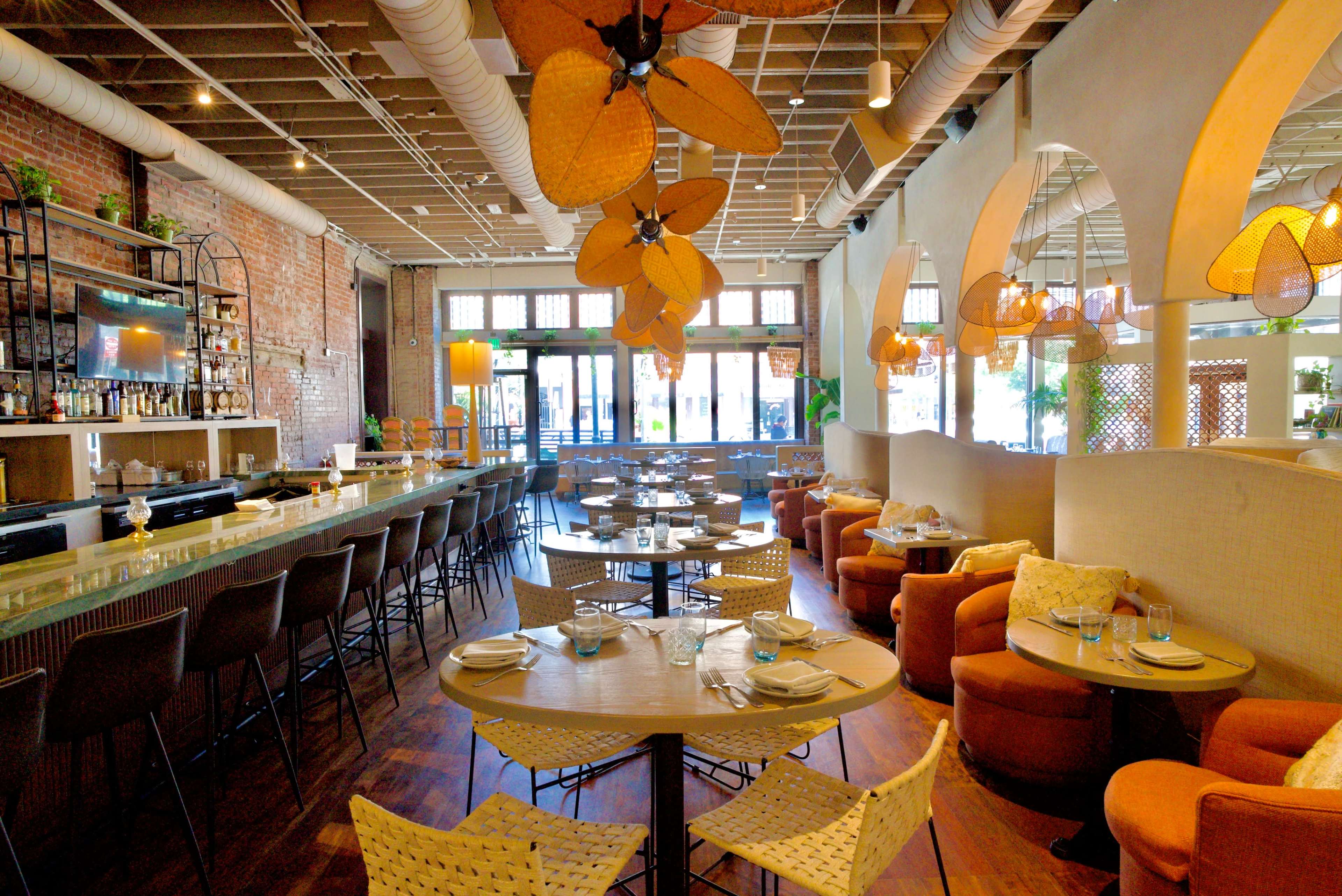The image shows a spacious restaurant interior featuring a bar area, round tables with woven chairs, and decorative light fixtures hanging from the ceiling.