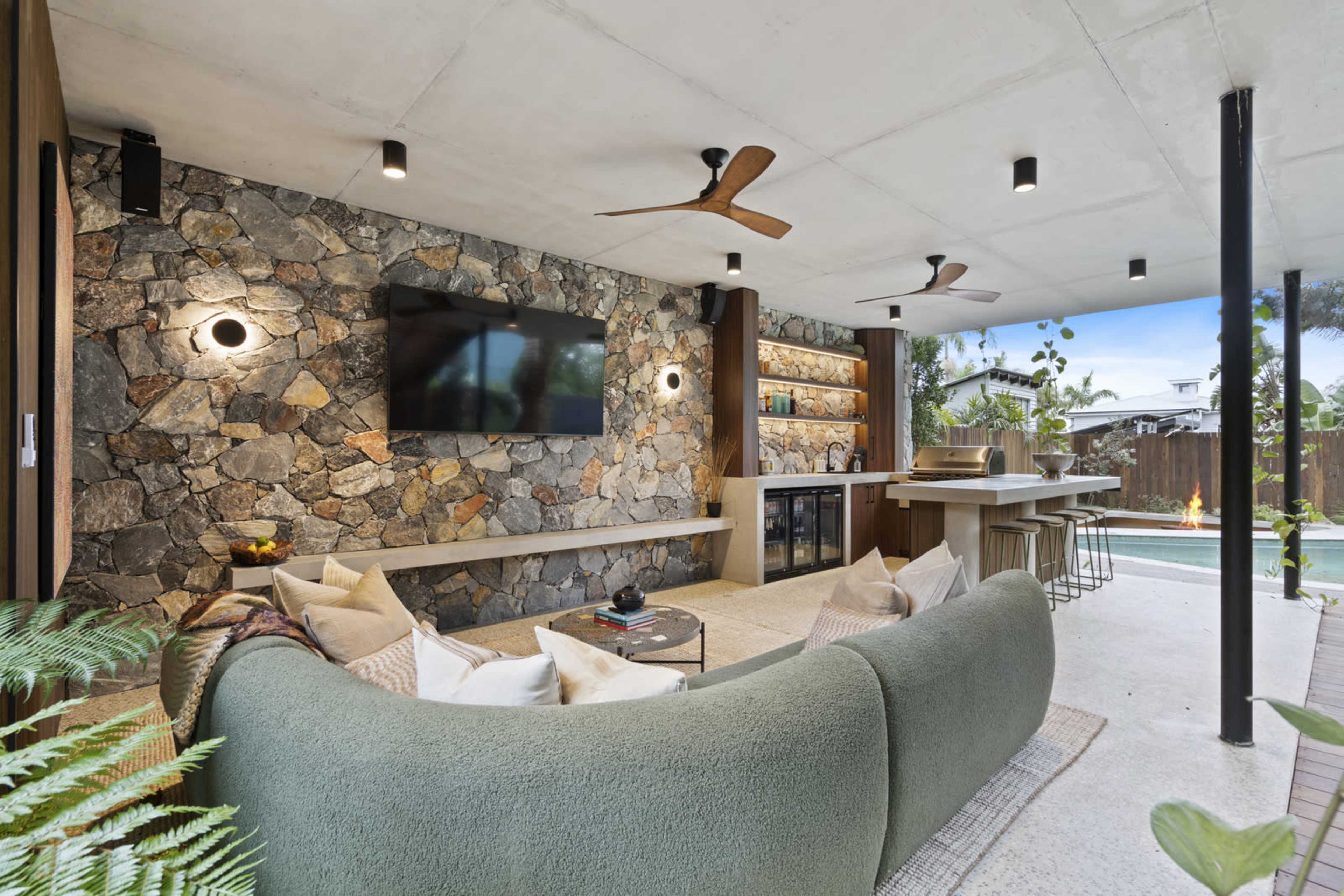 The image shows a modern outdoor living area with a curved green sofa, a stone accent wall featuring a TV, and a bar area next to a pool.