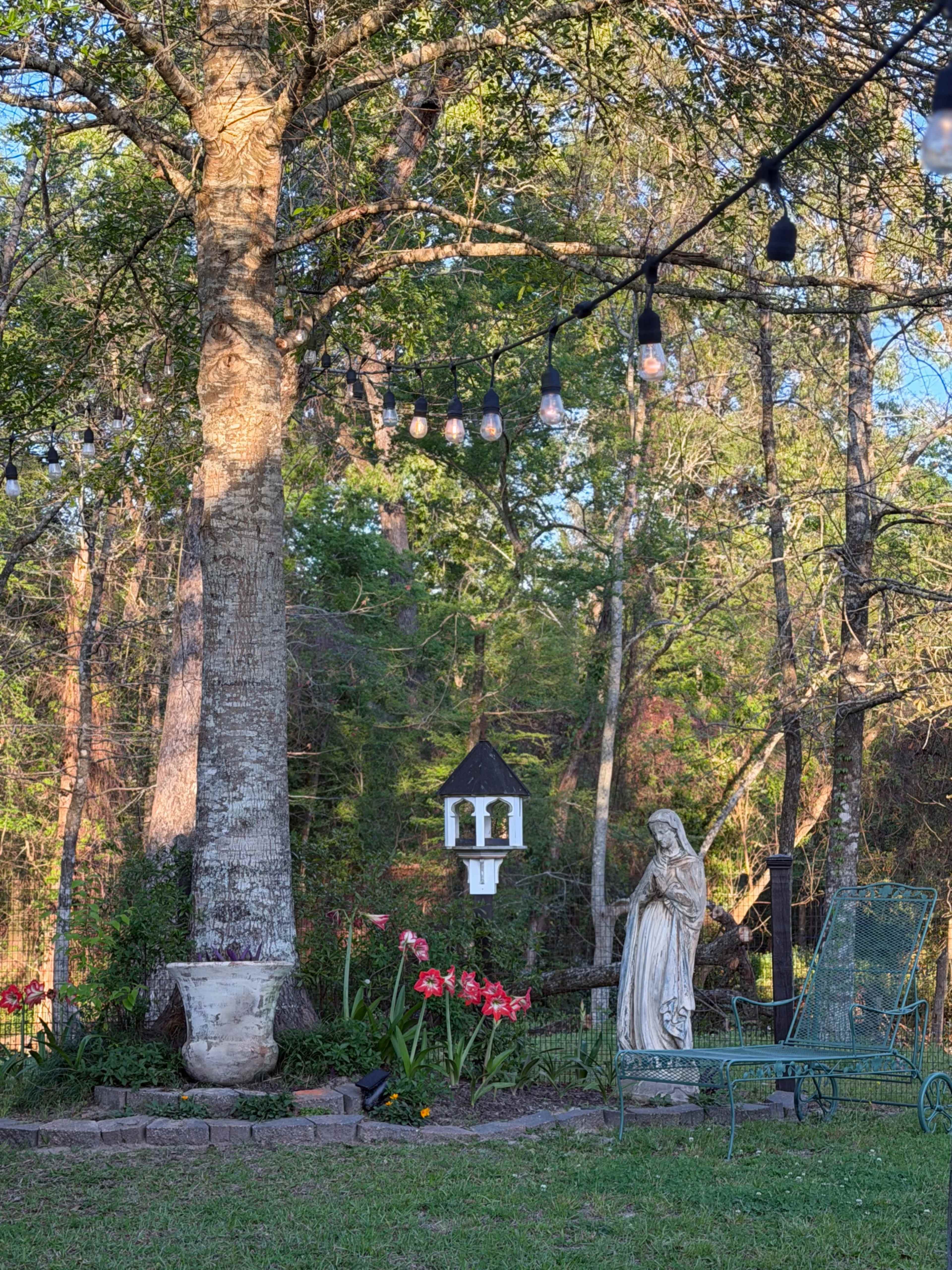 The image shows a garden scene with a large tree, a bird feeder hanging from a structure, a statue of a woman, and flowering plants surrounded by string lights.