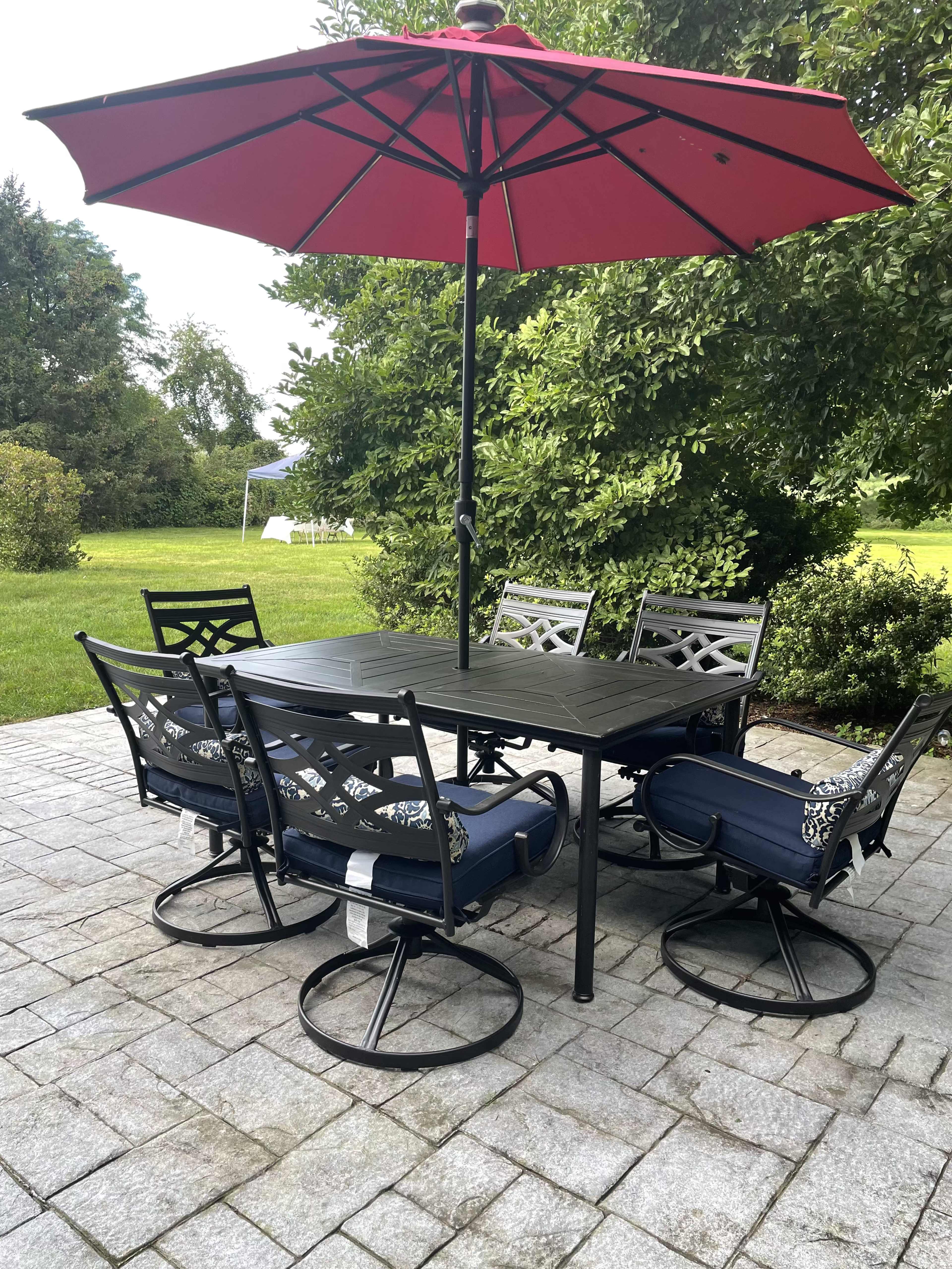 A patio table with six chairs and a red umbrella is set on a stone surface in a garden area.