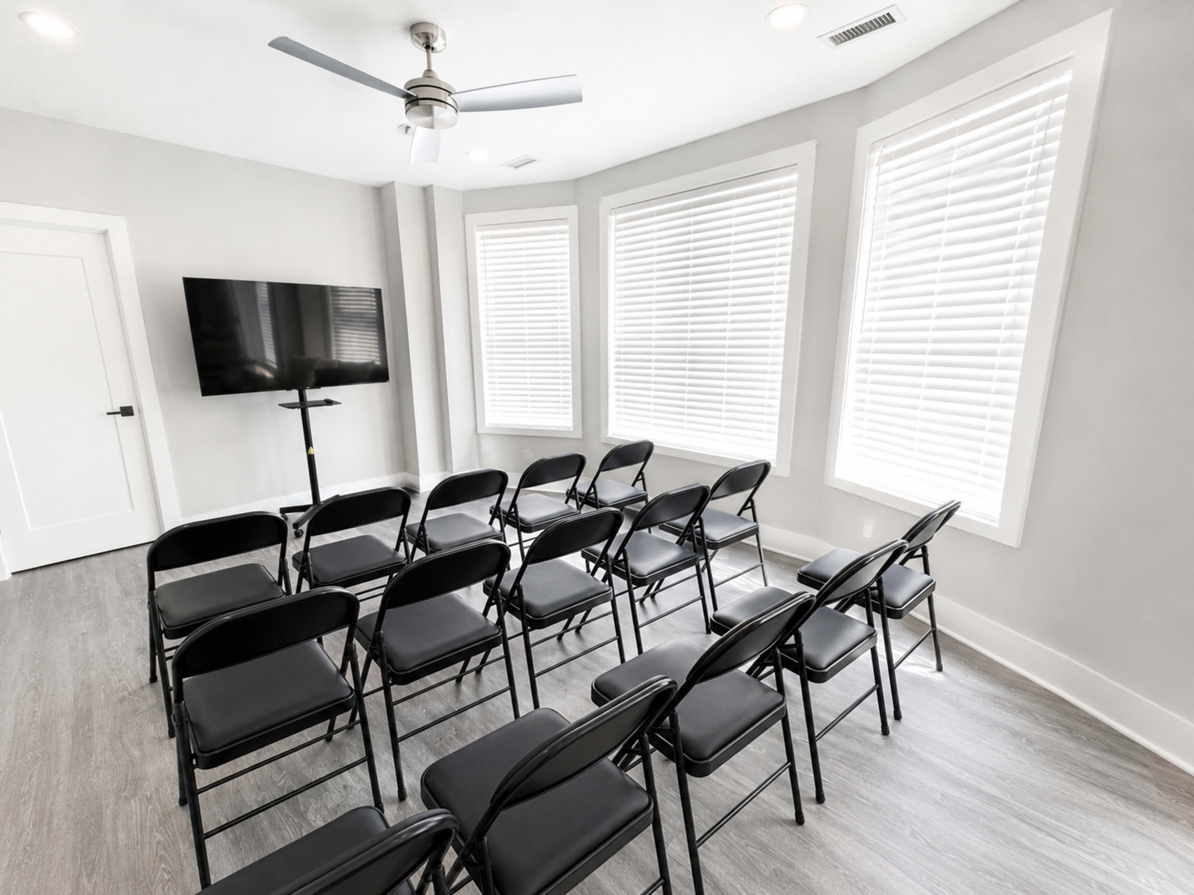 The image shows a lightly furnished meeting room with several rows of black folding chairs facing a wall-mounted television and large windows with blinds.