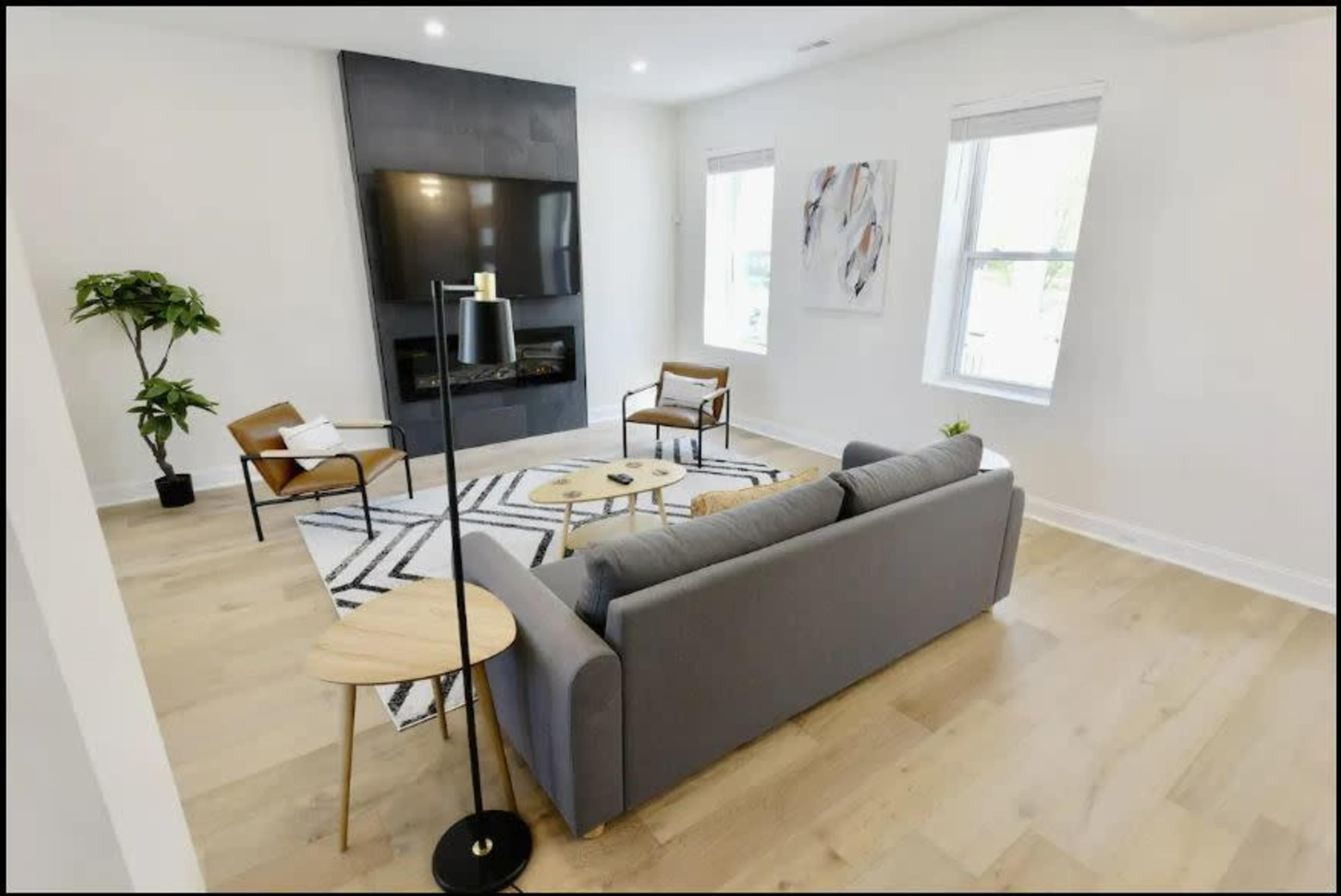 A modern living room features a gray sofa, two wooden chairs, a coffee table on a patterned rug, a wall-mounted television, and a potted plant.
