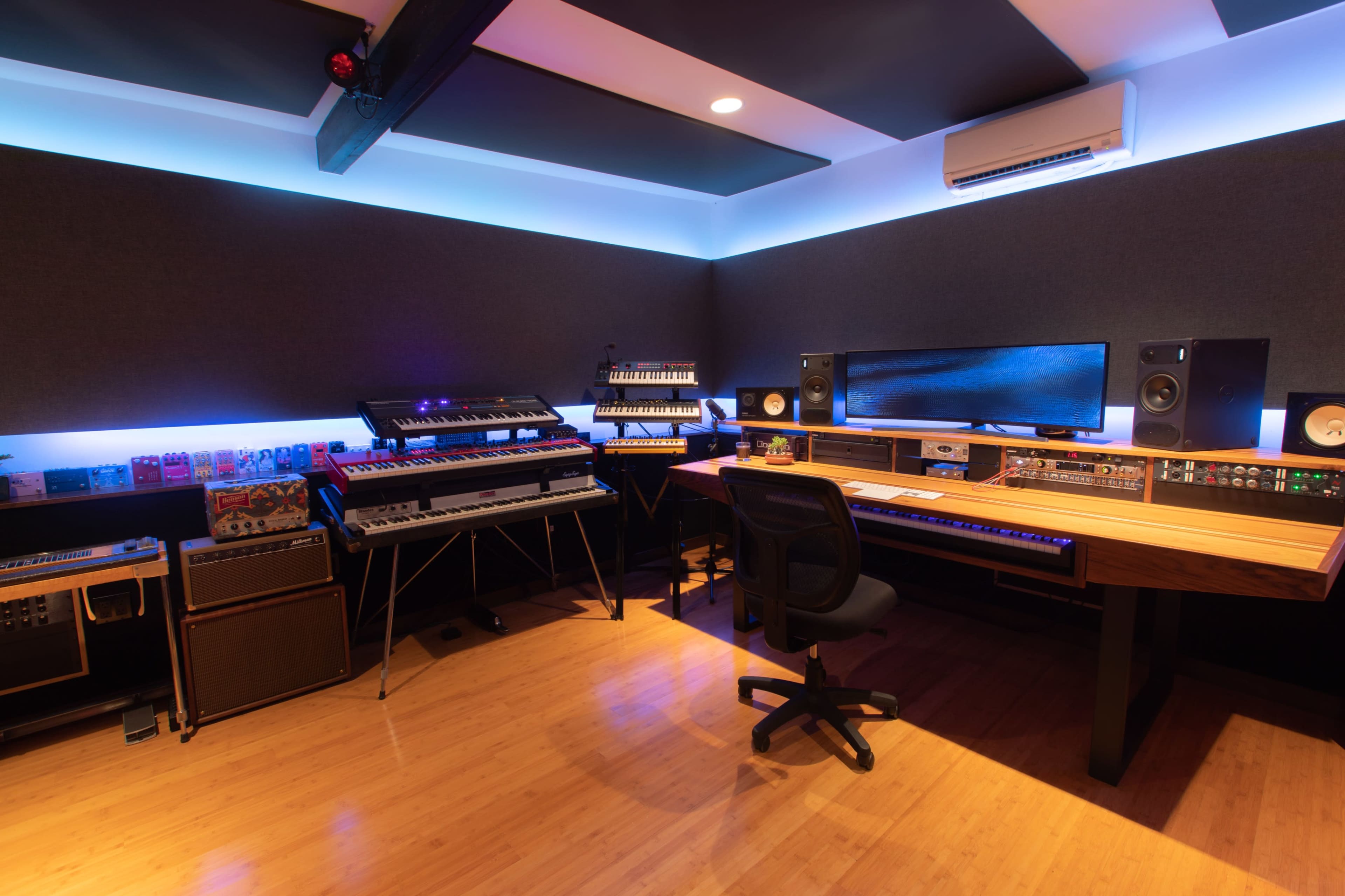 A modern music studio with various synthesizers, sound equipment, and a computer setup on a wooden desk, accented by blue LED lighting.