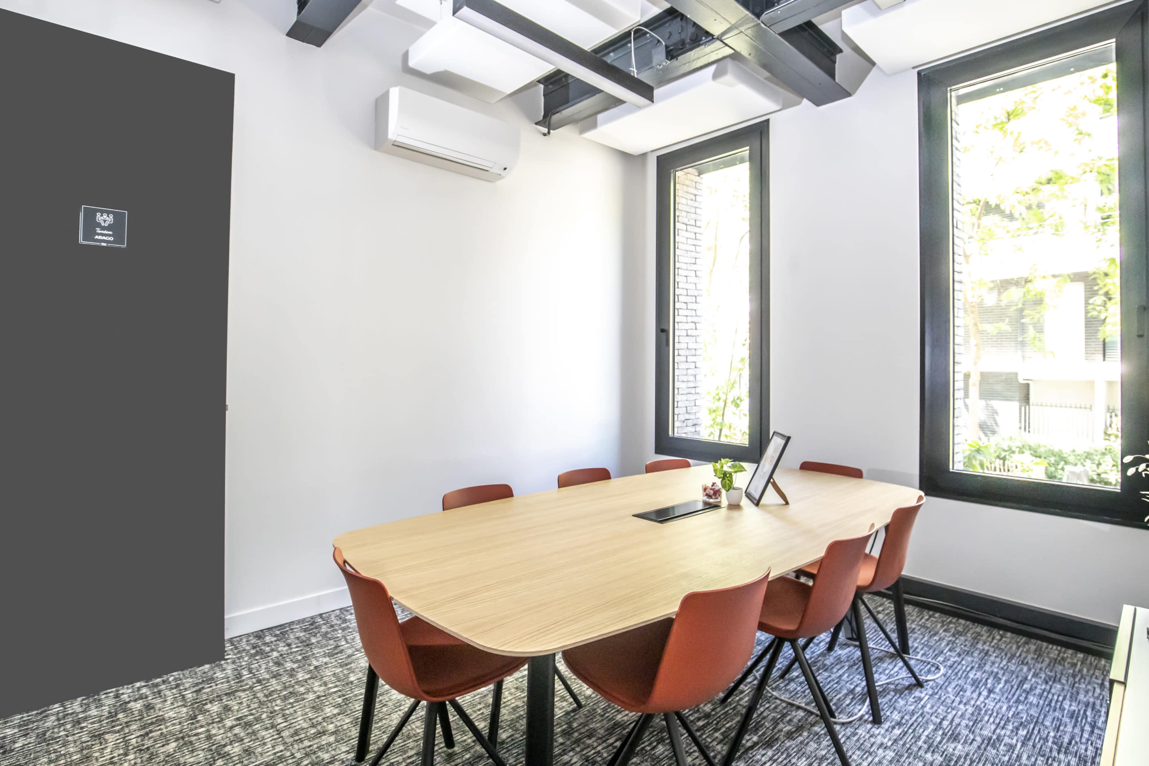 A modern meeting room with a wooden table surrounded by orange chairs and large windows letting in natural light.