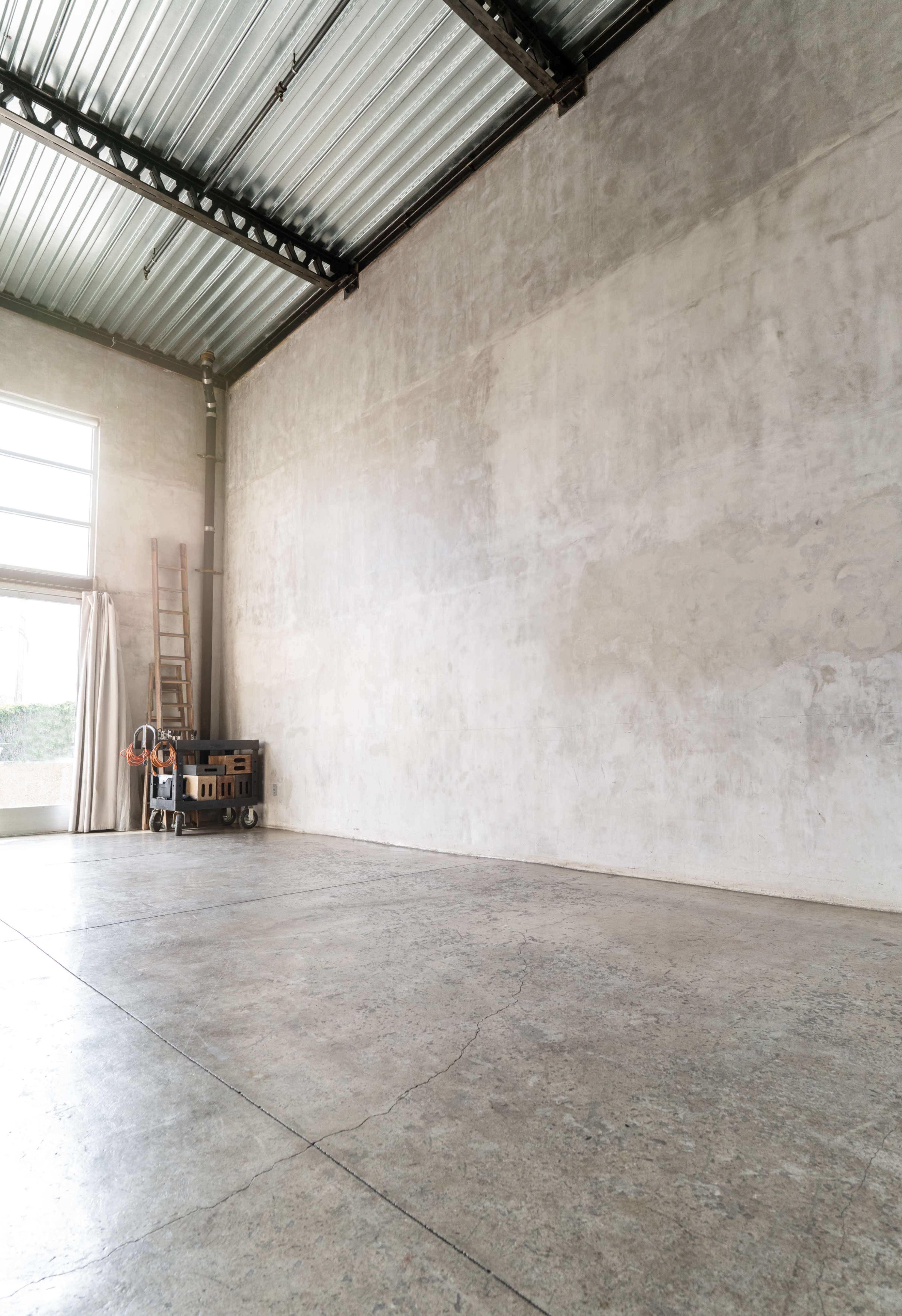 The image shows an empty industrial space with a concrete floor, a high ceiling, and a large window partially covered by a curtain.