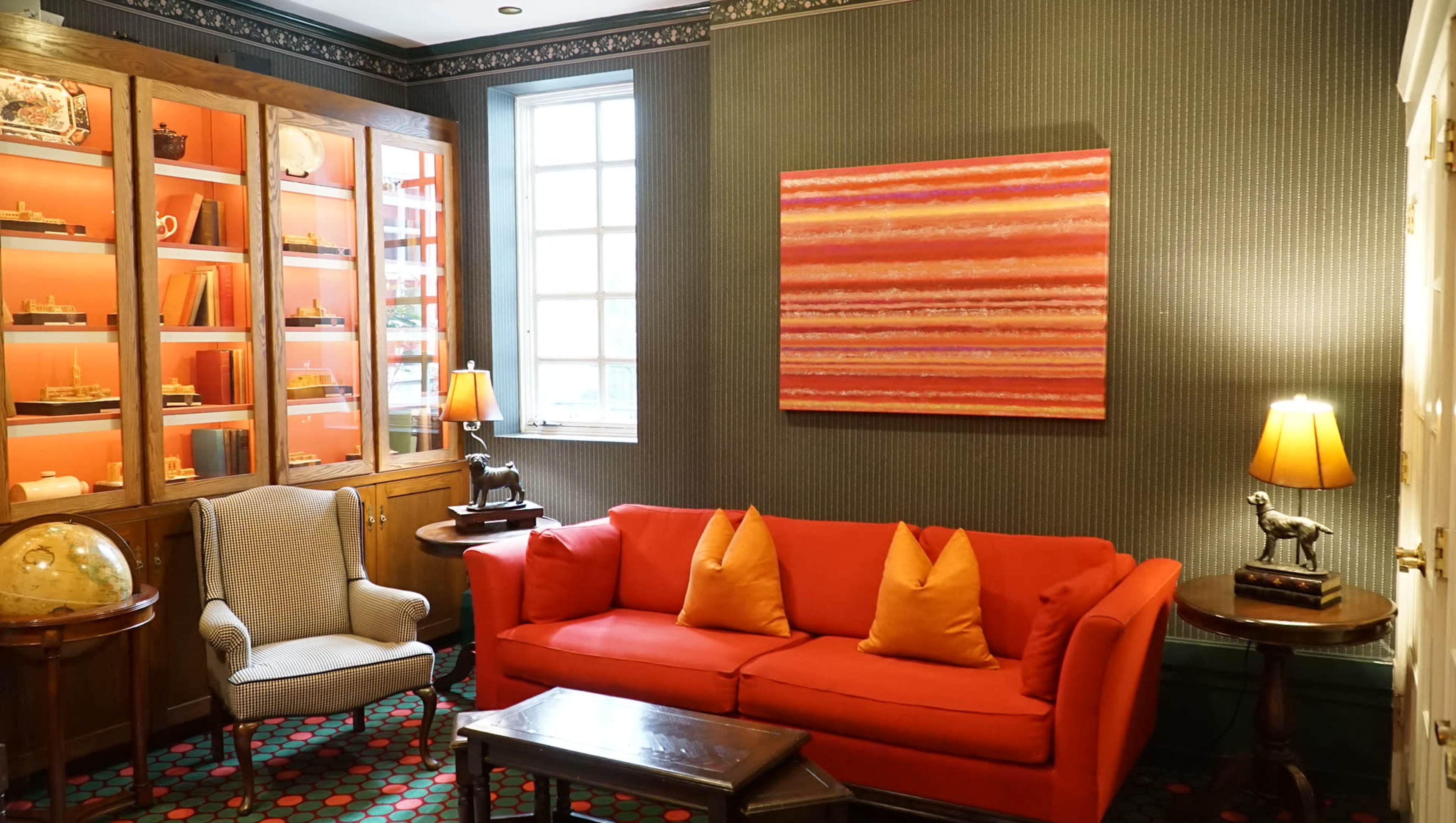 A cozy sitting area featuring an orange couch with decorative pillows, a coffee table, a globe, and illuminated side lamps, alongside a wall adorned with a colorful artwork and a display cabinet filled with various items.