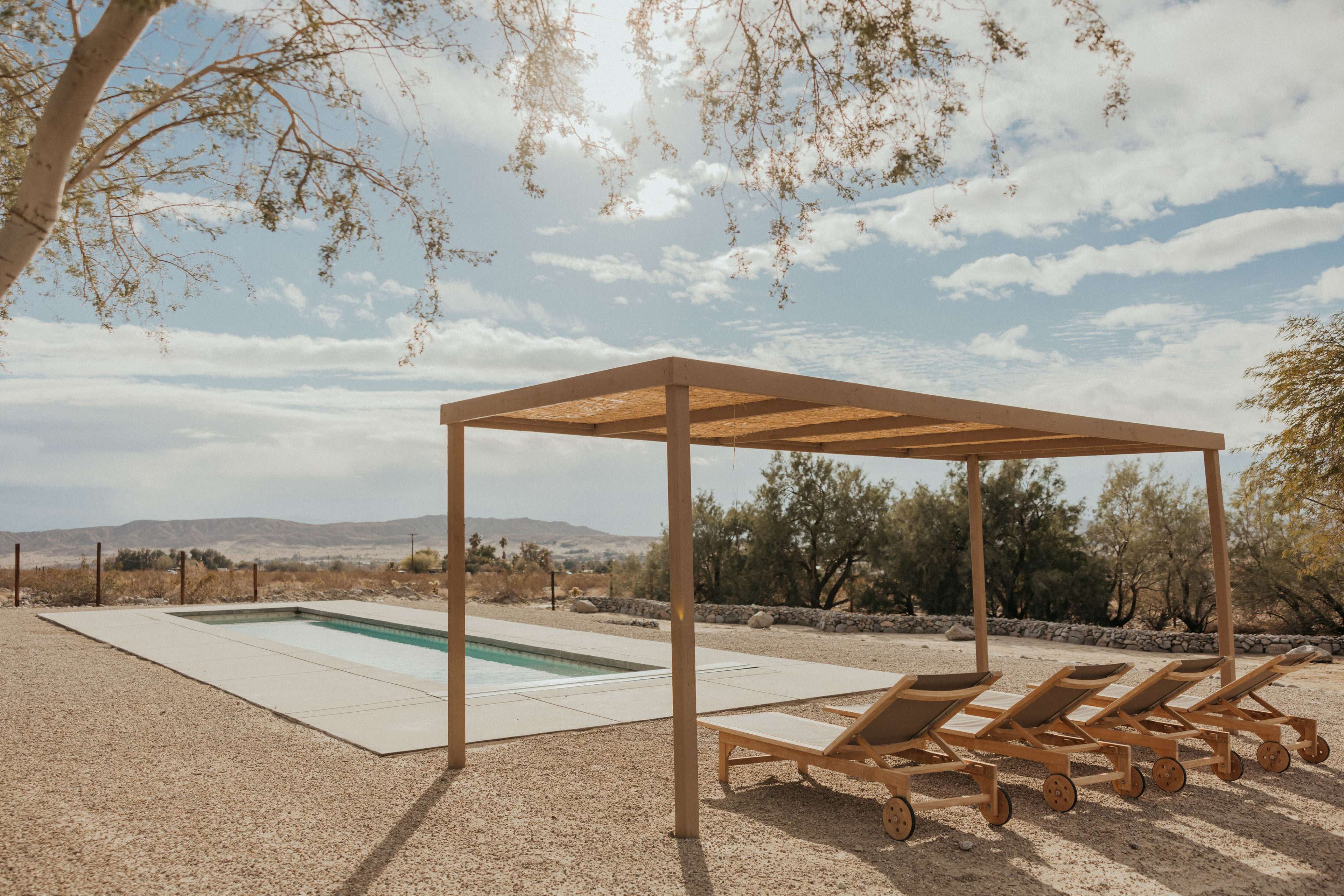 A simple wooden pergola shades a row of lounge chairs next to a rectangular swimming pool surrounded by a gravel landscape.