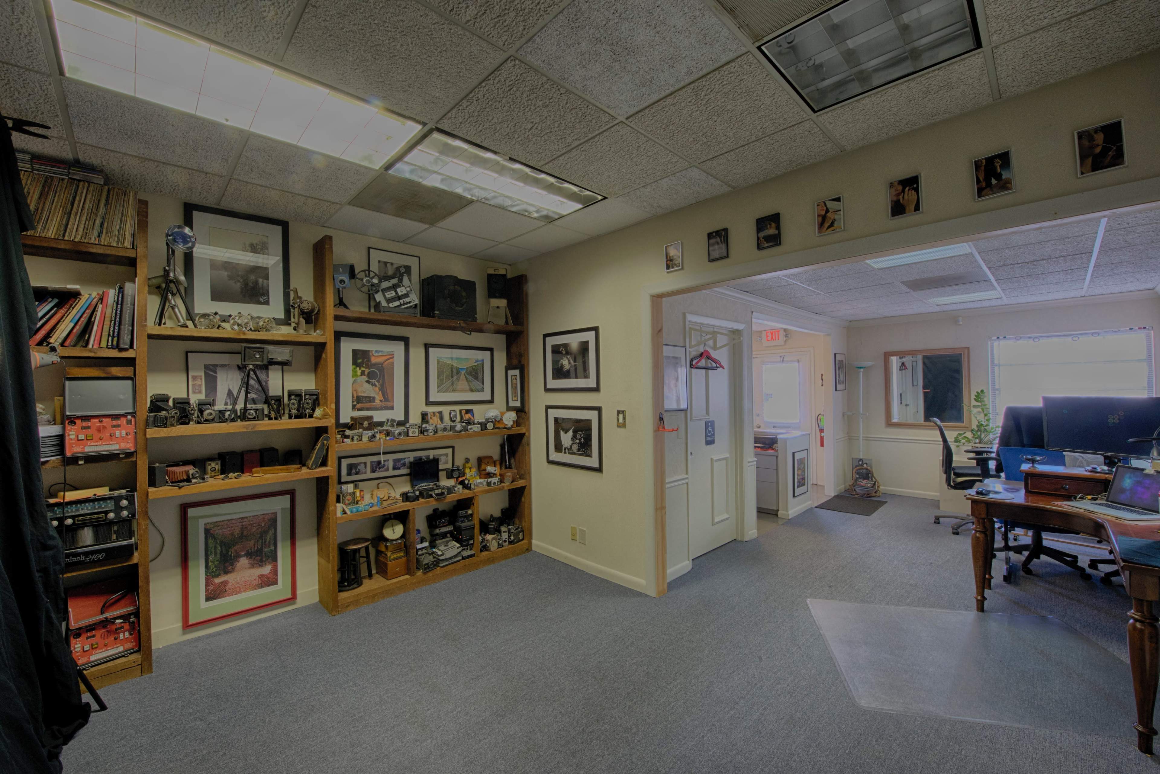 The image shows an office space with shelves displaying various cameras and framed photographs, leading to another room visible through an open door at the far end.